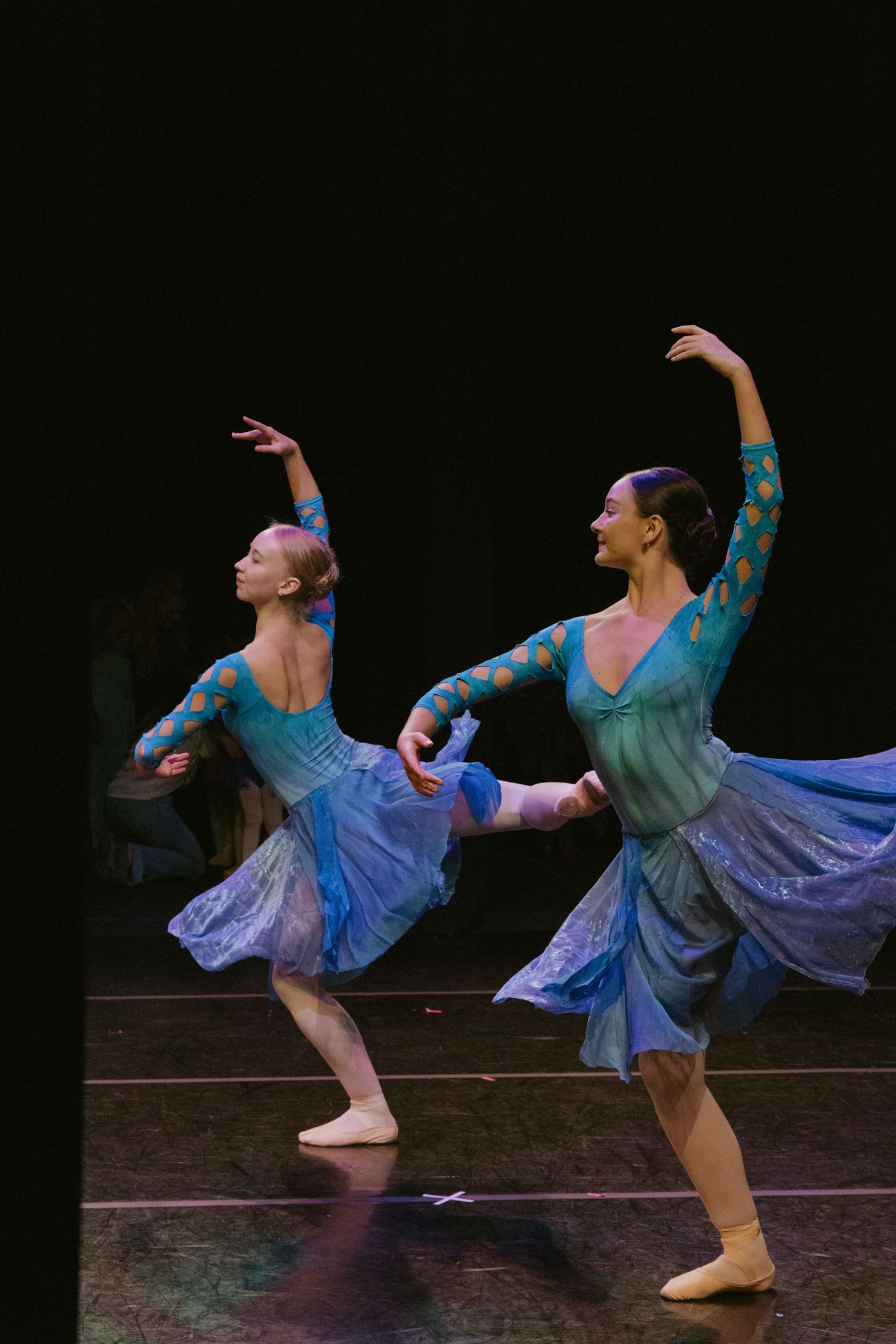 Two ballet dancers in flowing blue costumes perform synchronized movements against a dark background on stage.