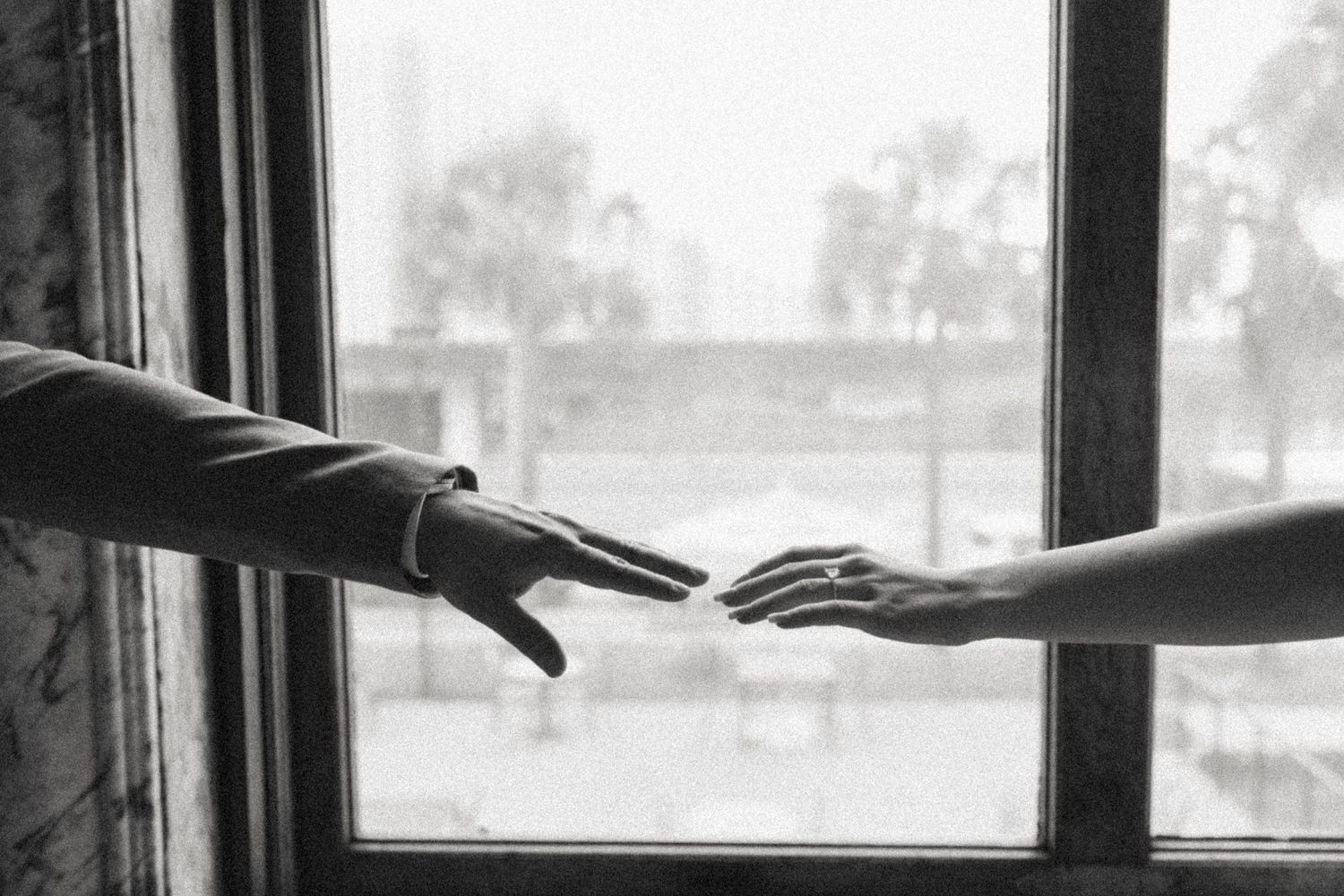 Black and white photo of two hands reaching toward each other by a window.