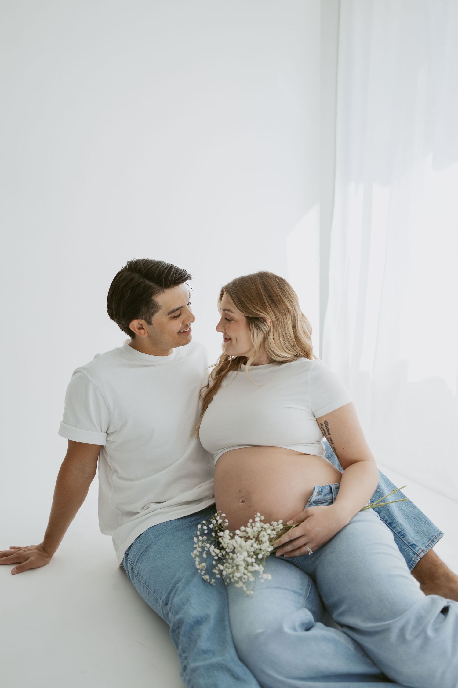Minimalist maternity photo with couple in white sitting against wall with baby's breath flowers.