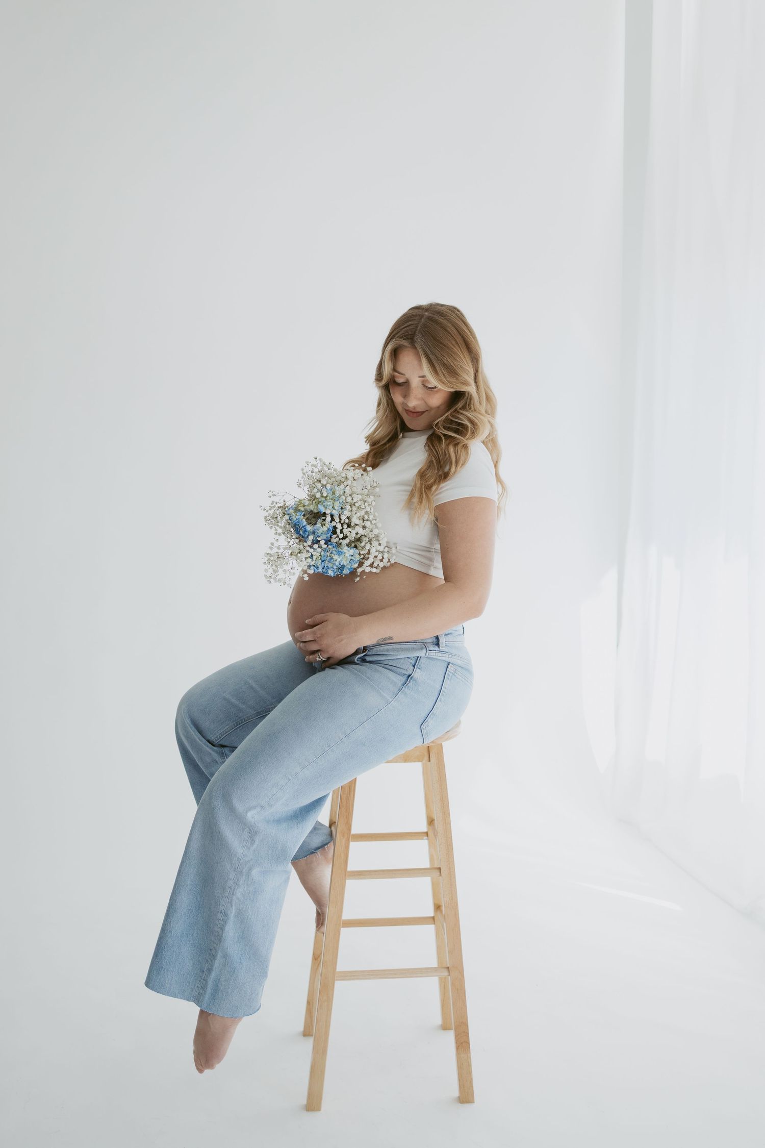 Person seated on wooden stool holding delicate blue flowers in a minimalist white setting.