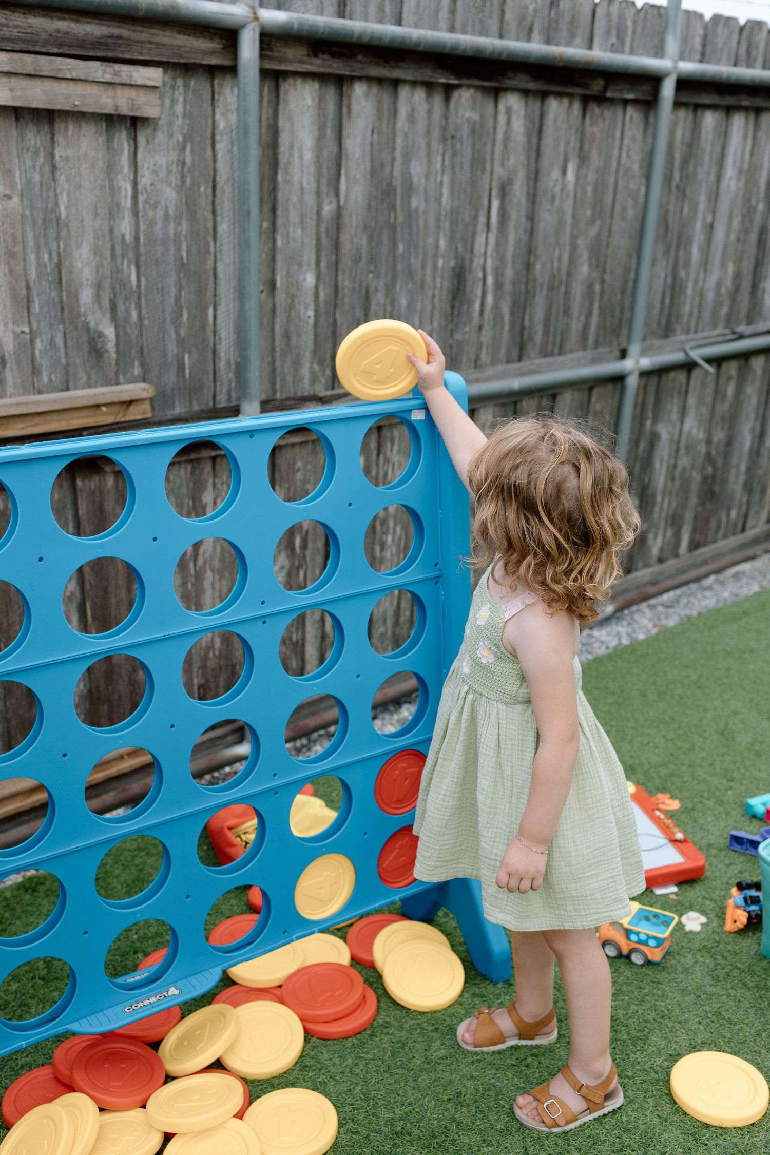 Child playing with giant outdoor Connect Four game on artificial turf with yellow and red game pieces.
