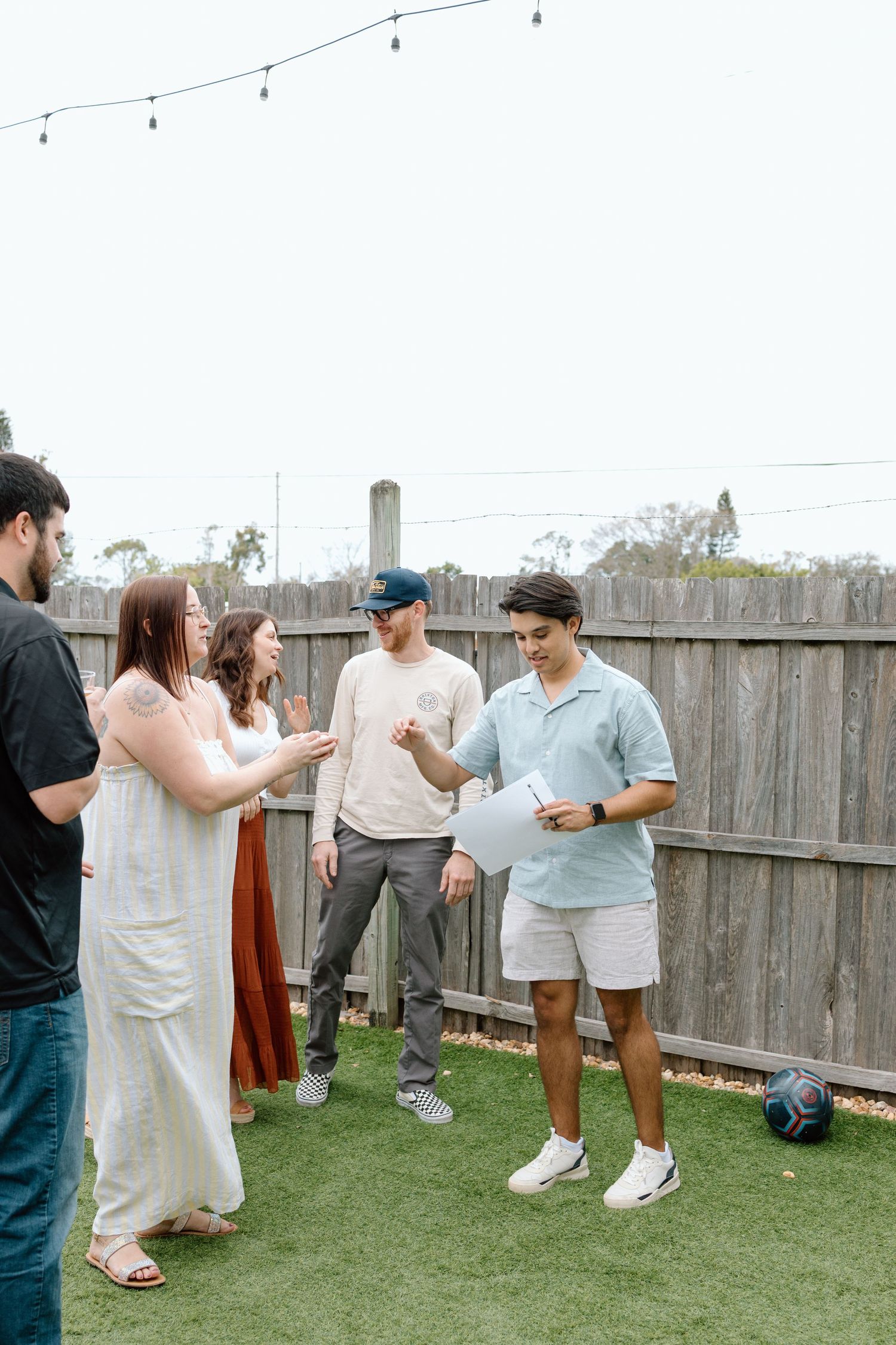 A group of people standing on artificial turf in a backyard having a casual gathering near a wooden fence with string lights.