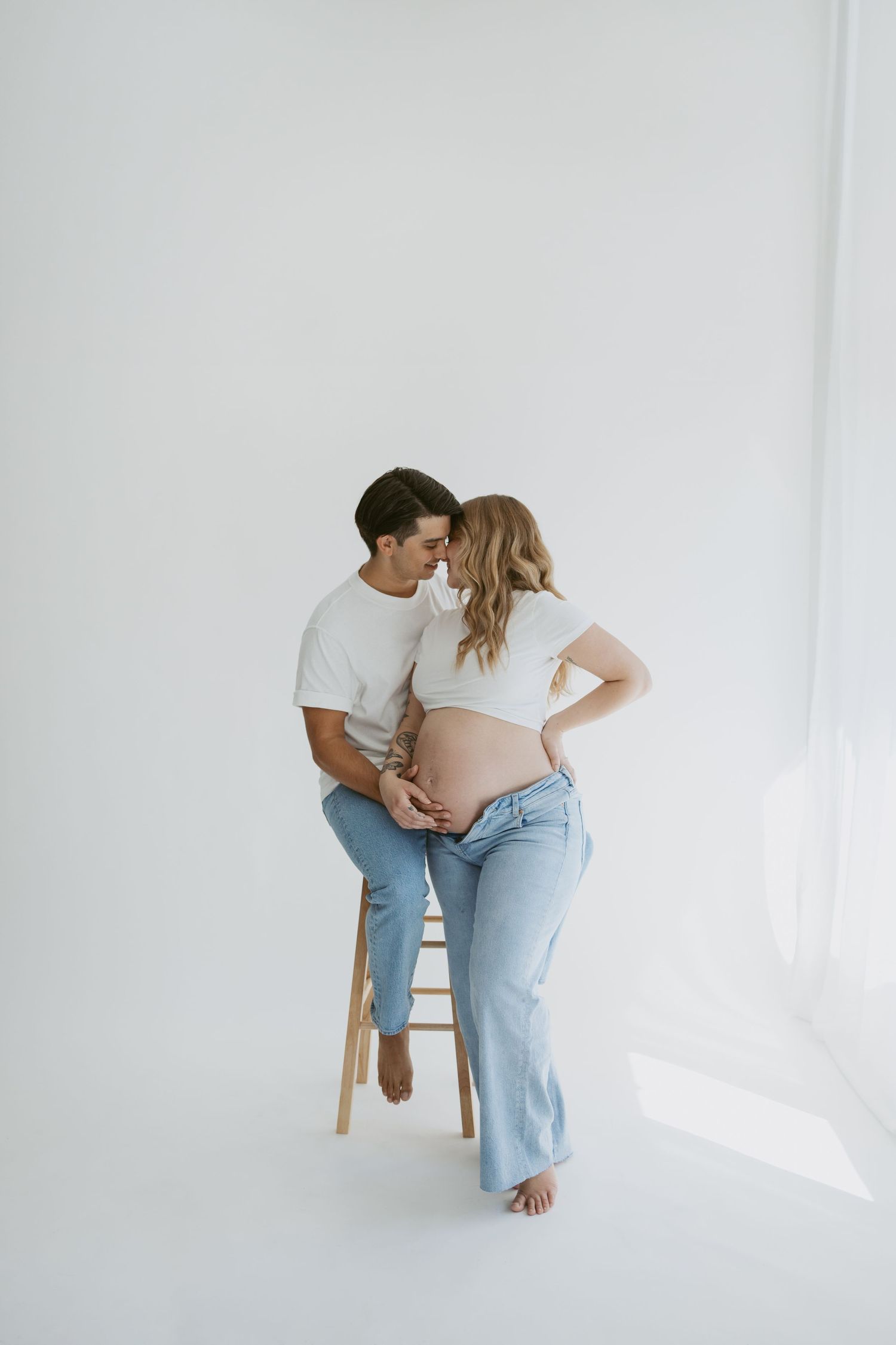 Artistic maternity portrait with couple sharing intimate moment on wooden stool.