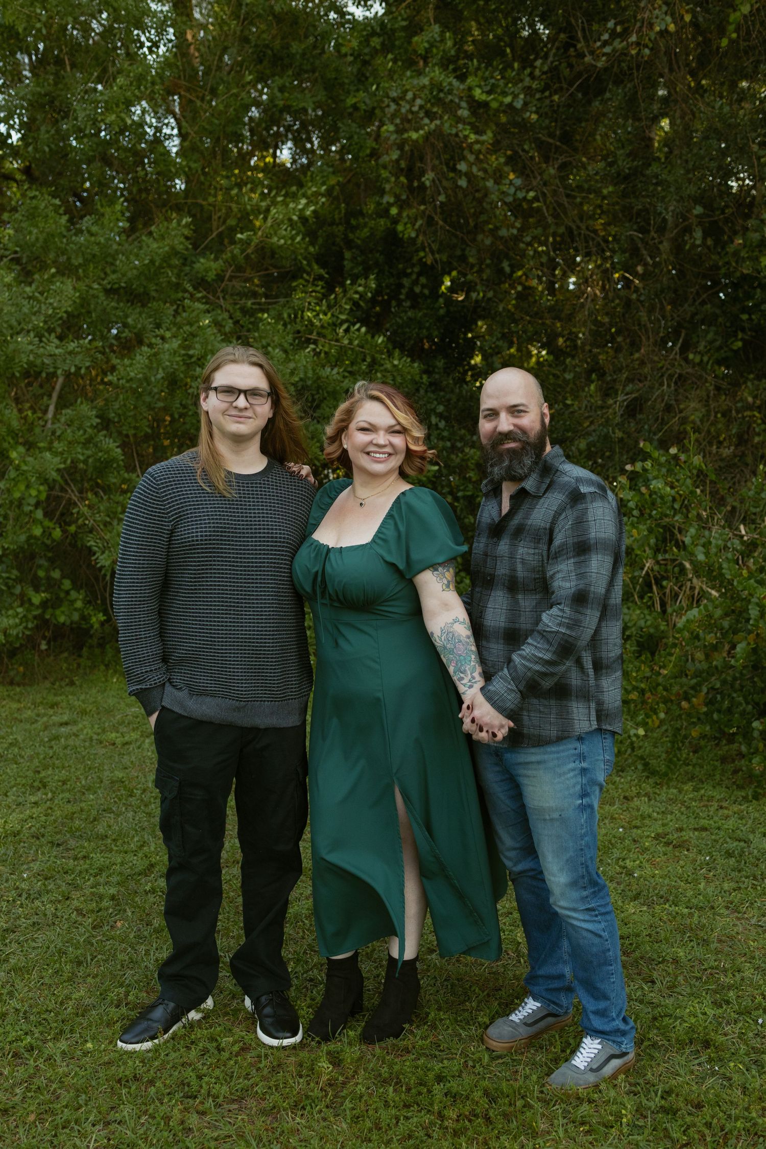 Outdoor family portrait featuring three people in casual attire against a green natural background.