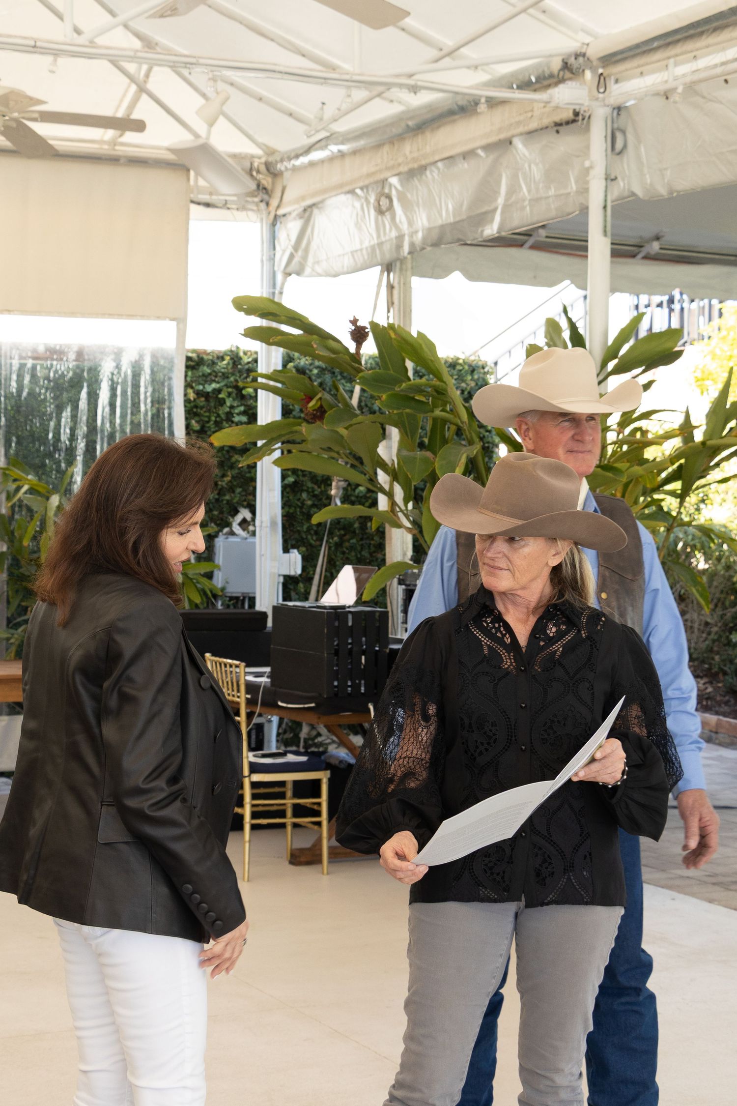 Three people having a discussion under a white canopy tent with tropical plants visible in the background.