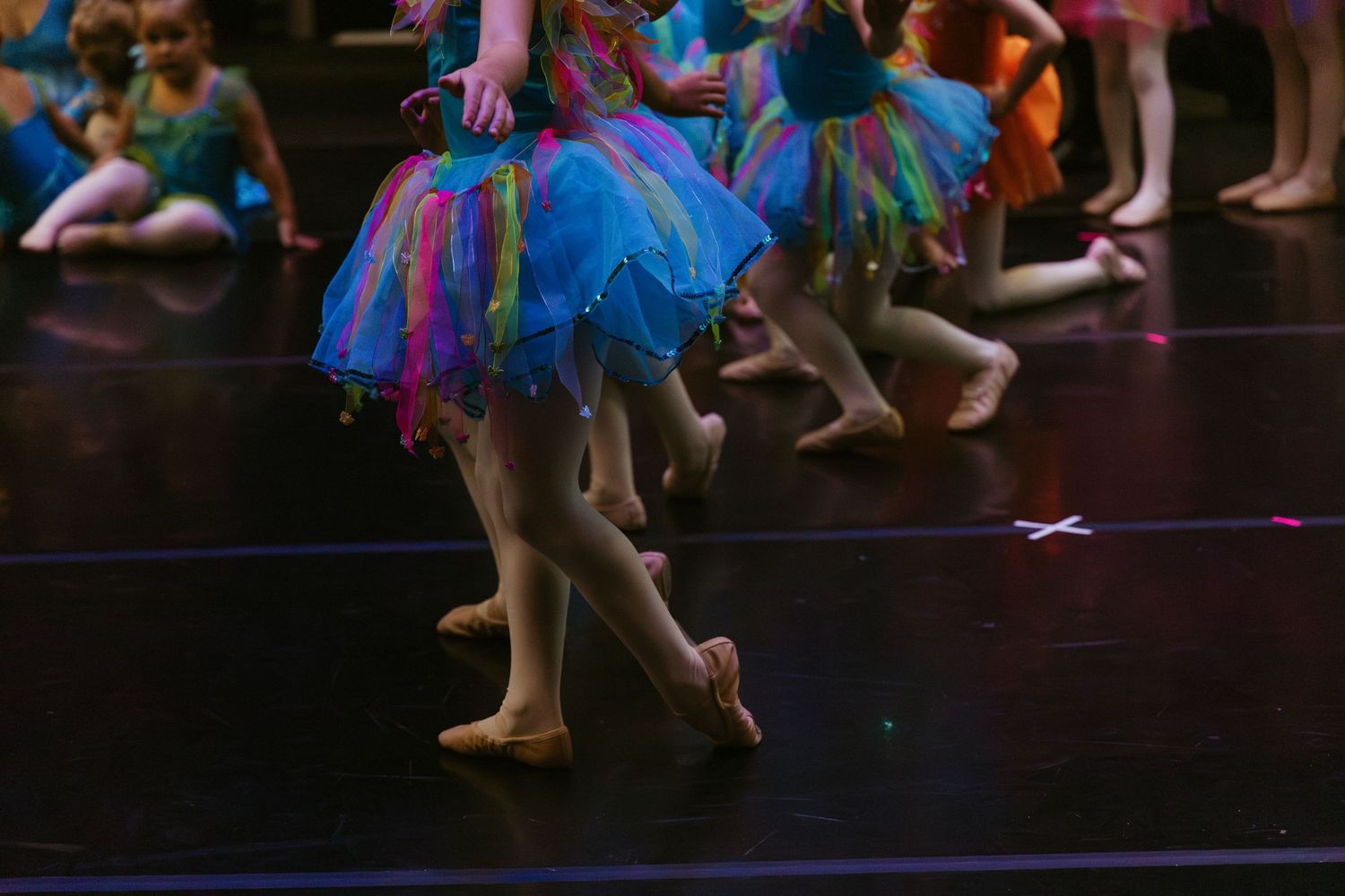 Young ballet dancers in colorful blue and pink tutus perform on stage with graceful footwork visible in silhouette.