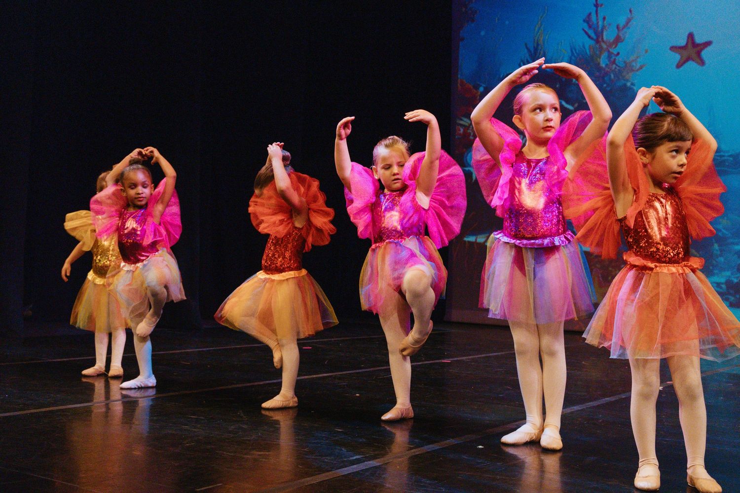 Dancers in bright pink and orange tutus perform synchronized movements on stage.