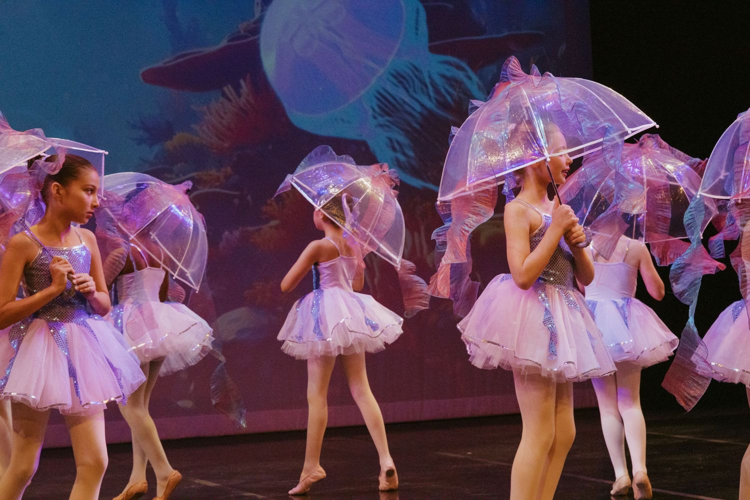 Ballet dancers in purple tutus pose with transparent umbrellas against blue stage lighting.