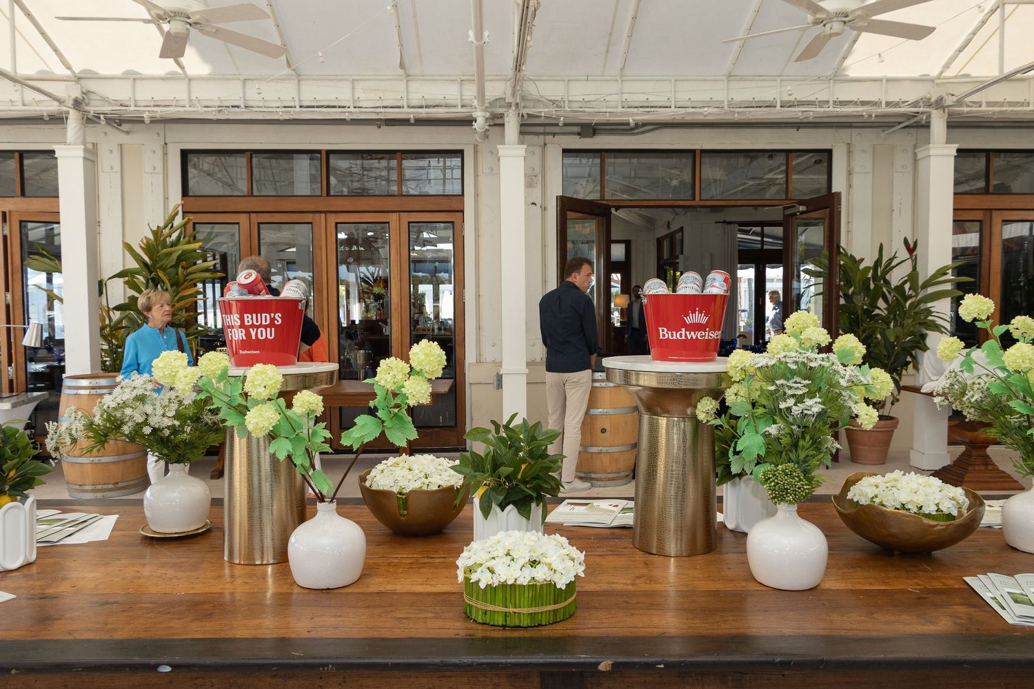 Elegant floral arrangements in white vases decorate a wooden table at an event with windows and greenery in background.