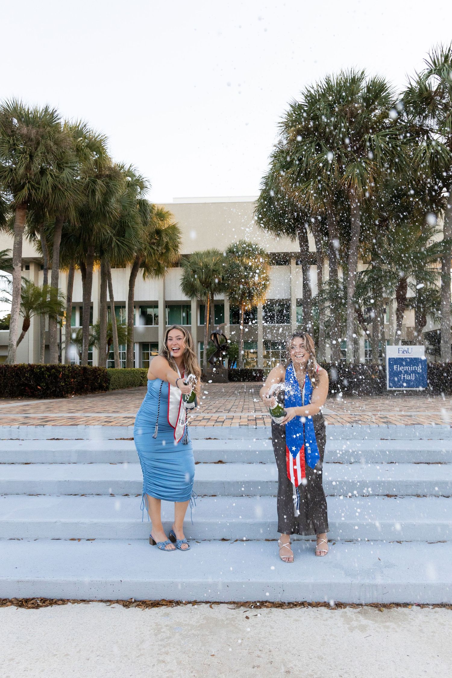 Two people celebrating on steps with confetti and champagne in front of a building with palm trees.