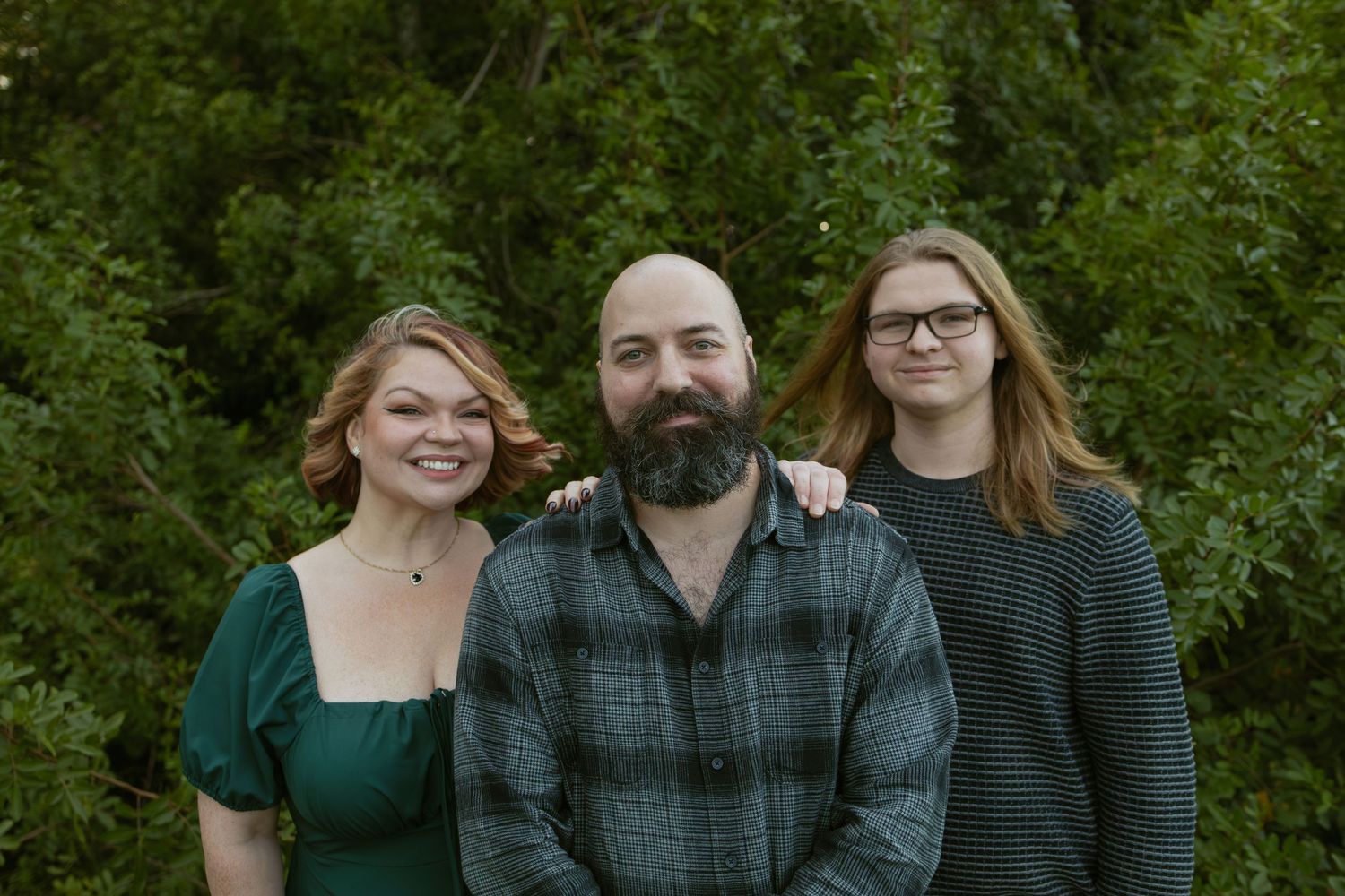 Three friends pose casually together against a leafy green background.
