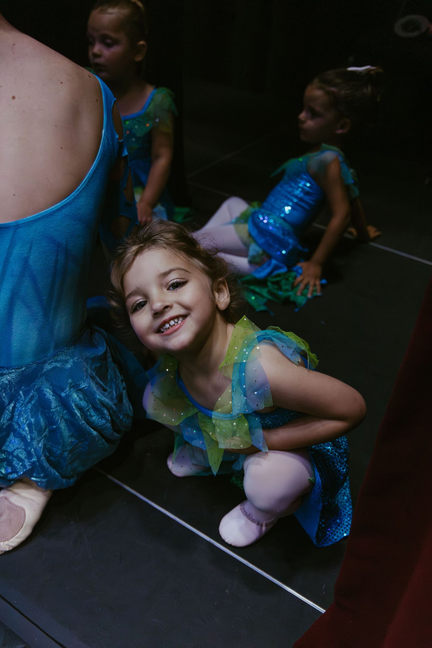 Young dancers in sparkly blue and green performance costumes wait backstage at a dance recital.