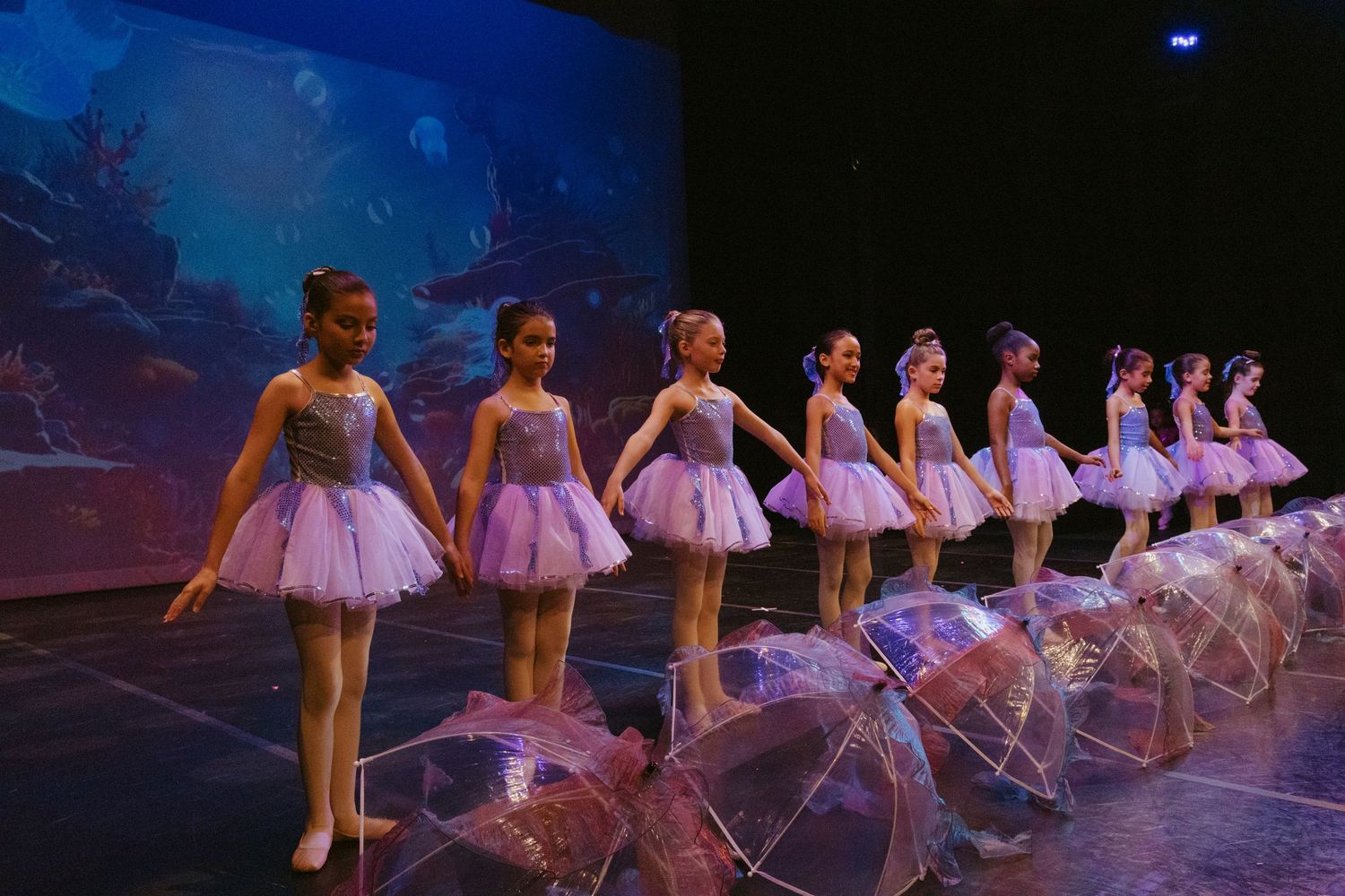 Ballet dancers in lavender tutus pose with clear umbrellas on a dimly lit stage.