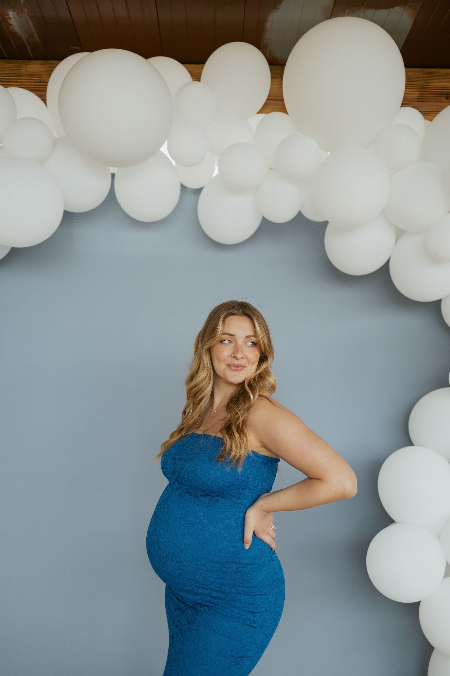 Expectant woman in a blue fitted dress poses beneath an elegant white balloon arch against a gray backdrop.