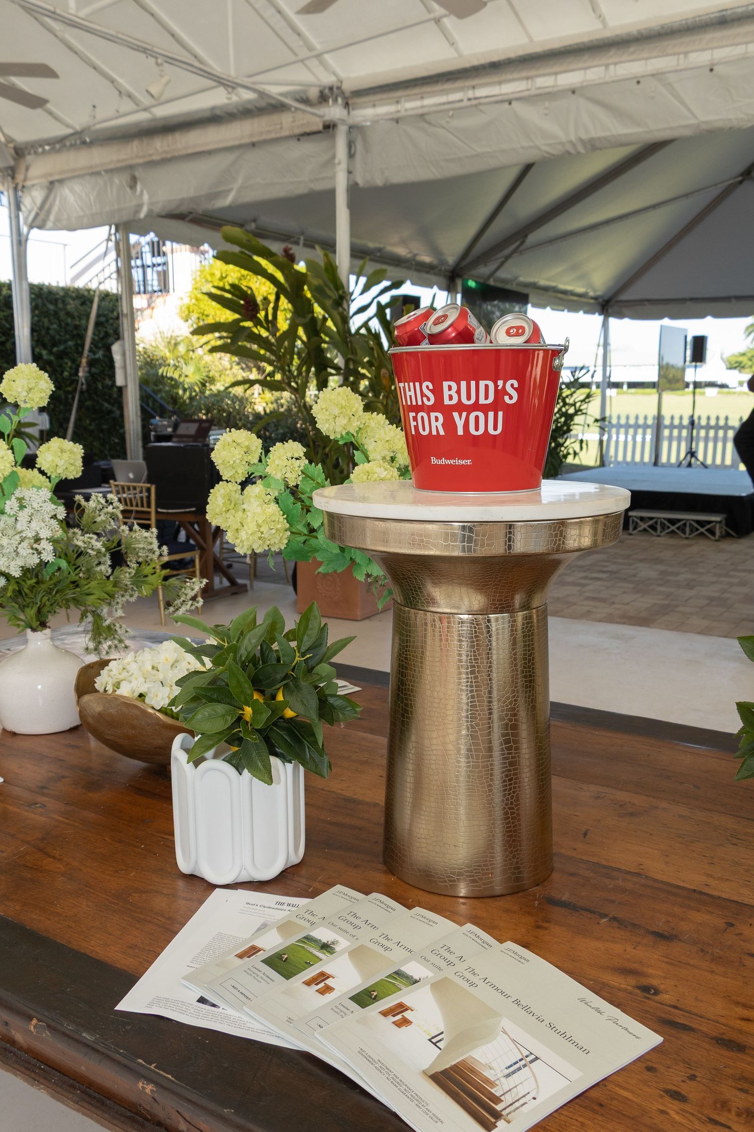 Red Budweiser bucket displayed on gold pedestal with white floral arrangements and printed materials on wooden table under tent.