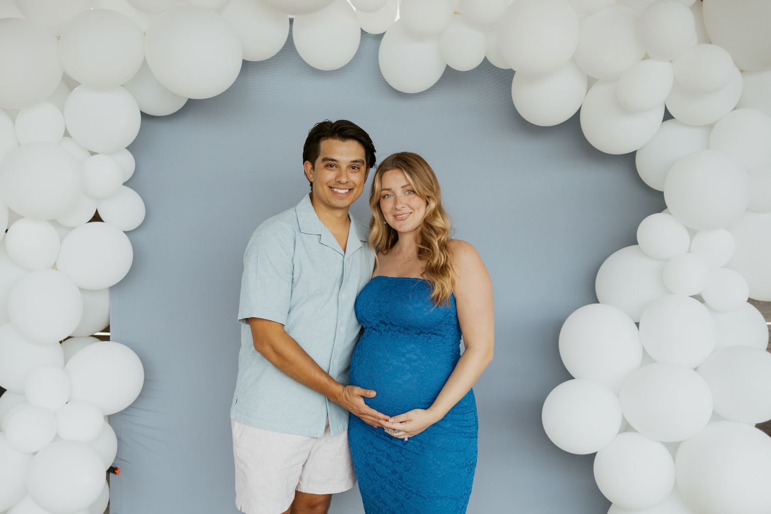 A couple poses under a white balloon arch against a blue backdrop, with one wearing a blue dress and another in white shorts.