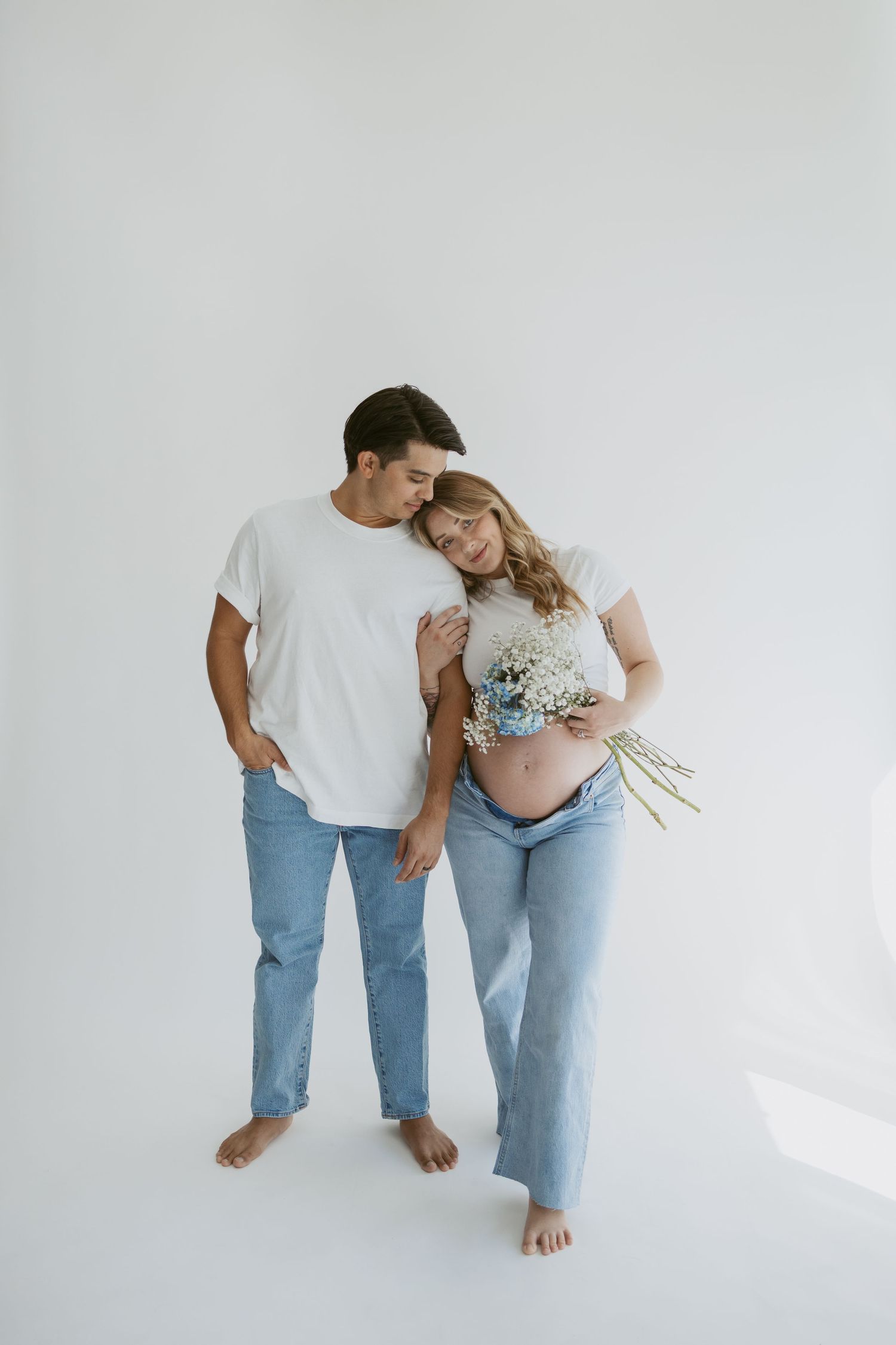 Expecting couple in casual attire holding baby's breath flowers against white backdrop.
