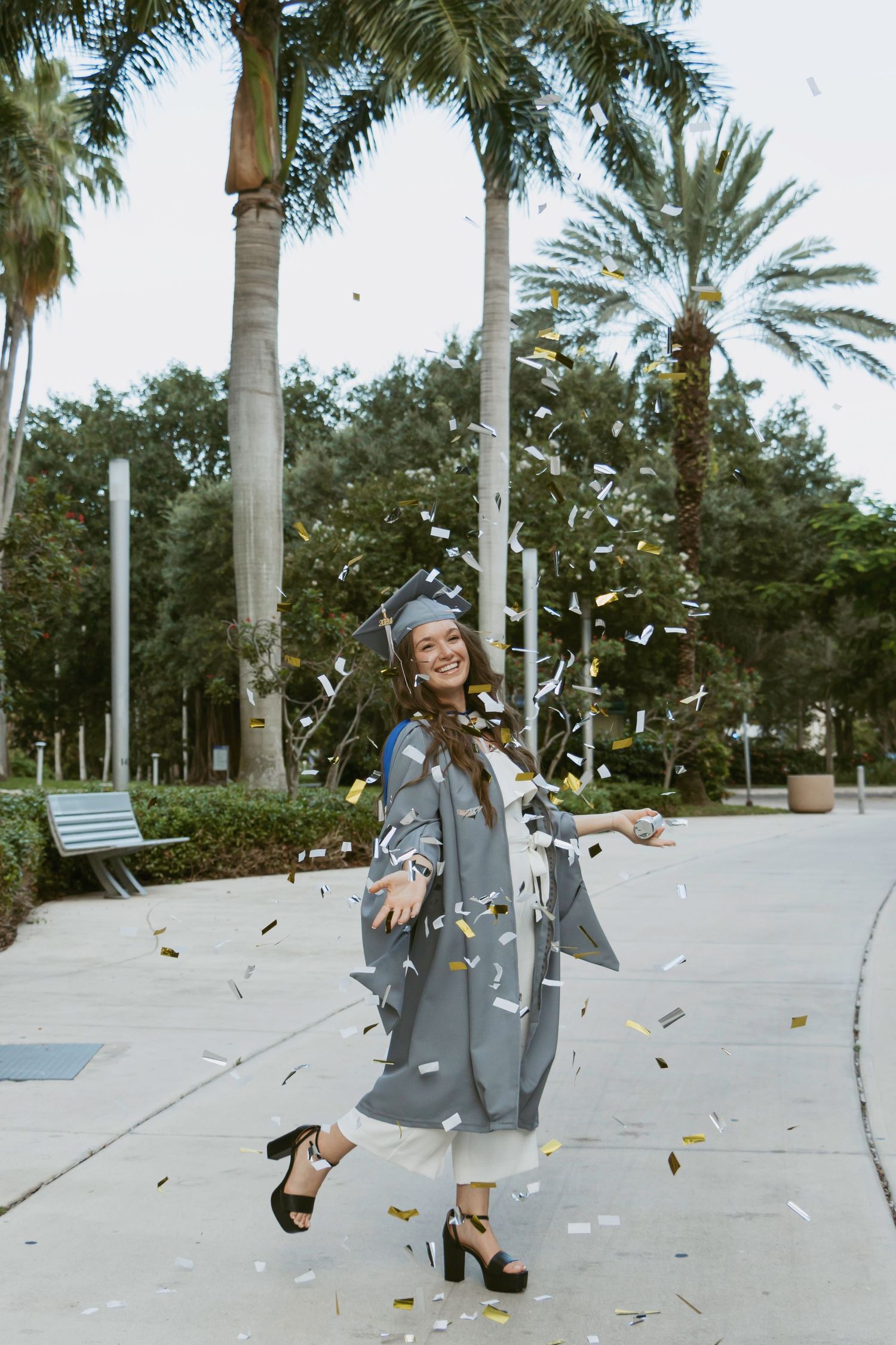 Graduate in grey graduation gown twirls joyfully under palm trees as confetti falls around her.
