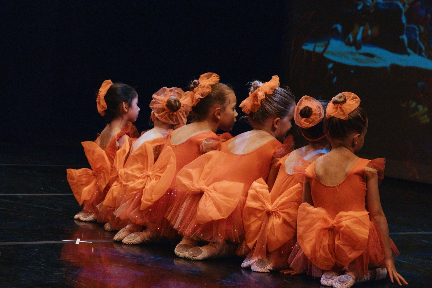 Line of dancers in matching orange dresses with bows perform synchronized movements.