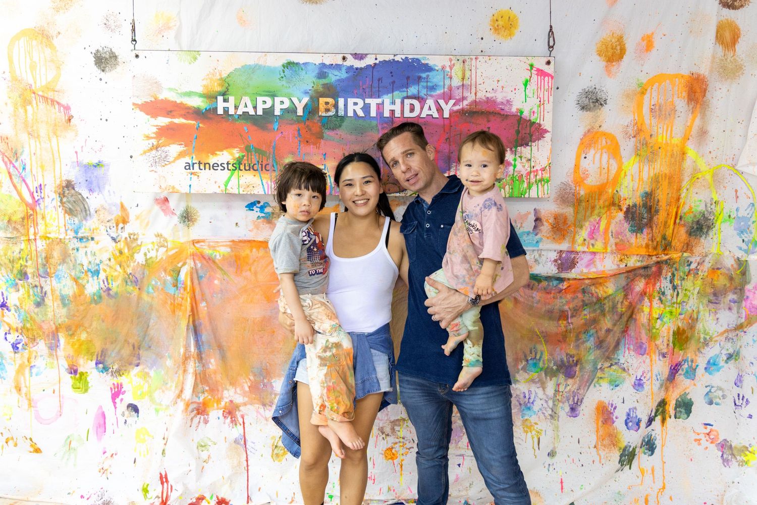 A family poses together against a colorful paint splattered wall with 'Happy Birthday' text at a celebration.