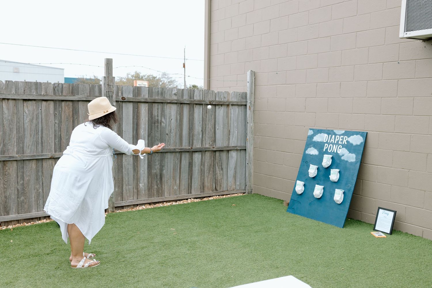 Person in white dress throws object at blue game board on artificial turf in backyard setting.