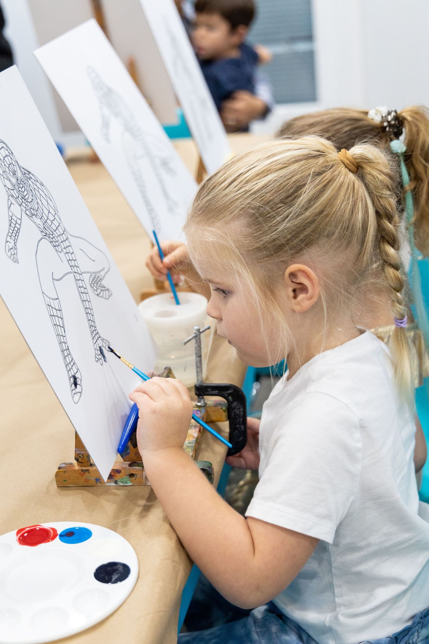 Young student in white shirt concentrates while drawing at an art easel with watercolor paints on desk.