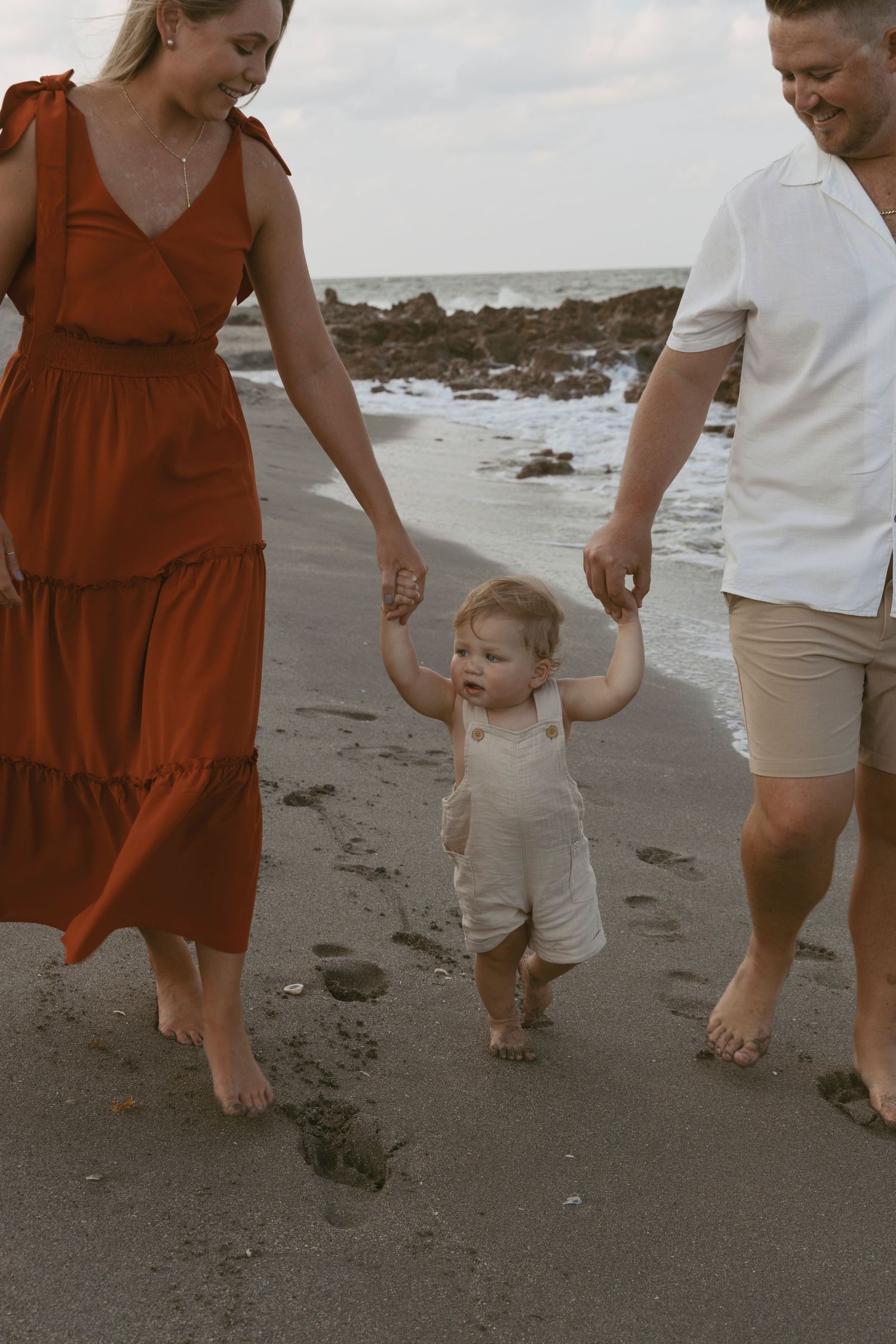 Parents holding hands with toddler while walking barefoot on sandy beach.