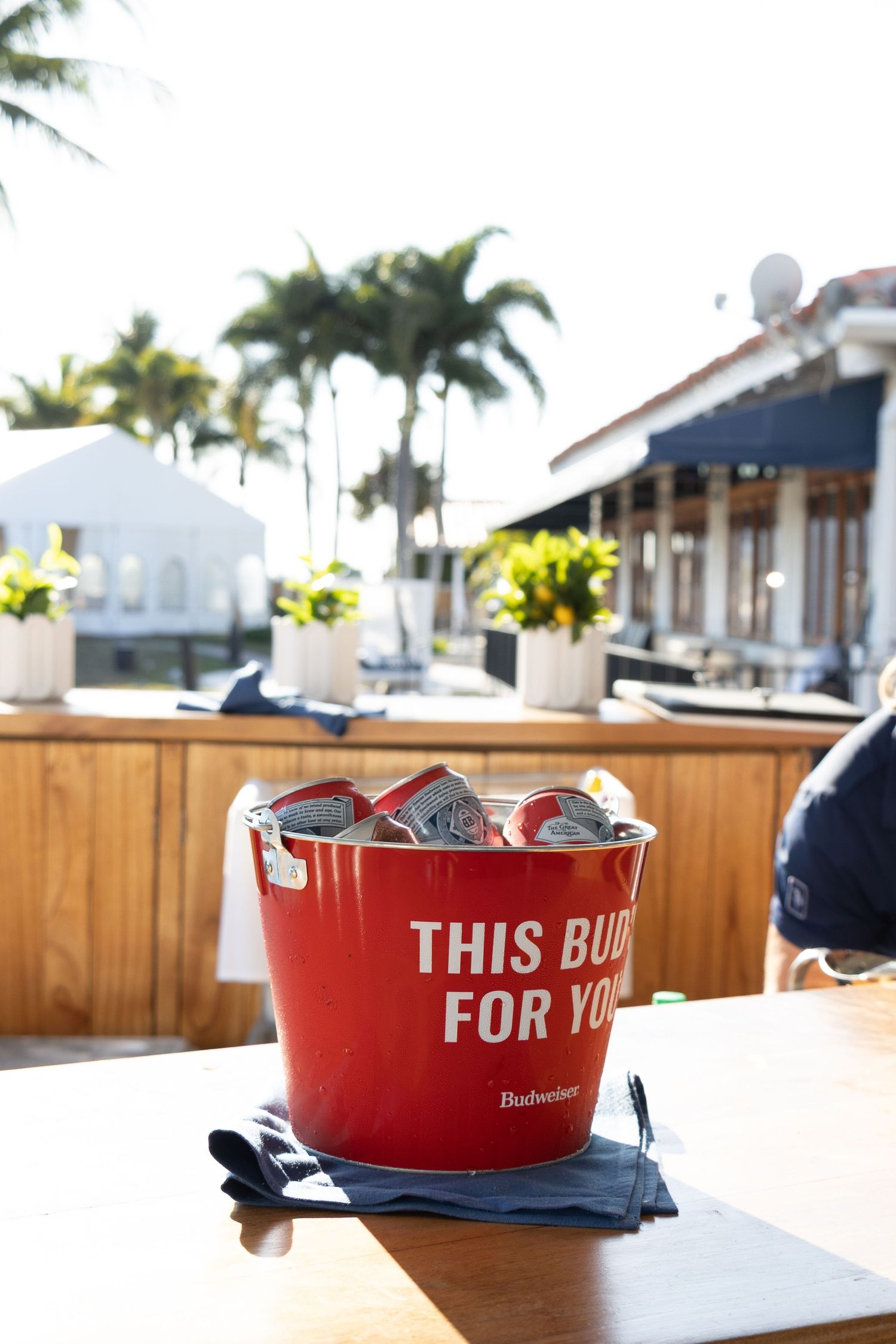 A red Budweiser beer bucket filled with ice and drinks sits on an outdoor patio table with palm trees in the background.