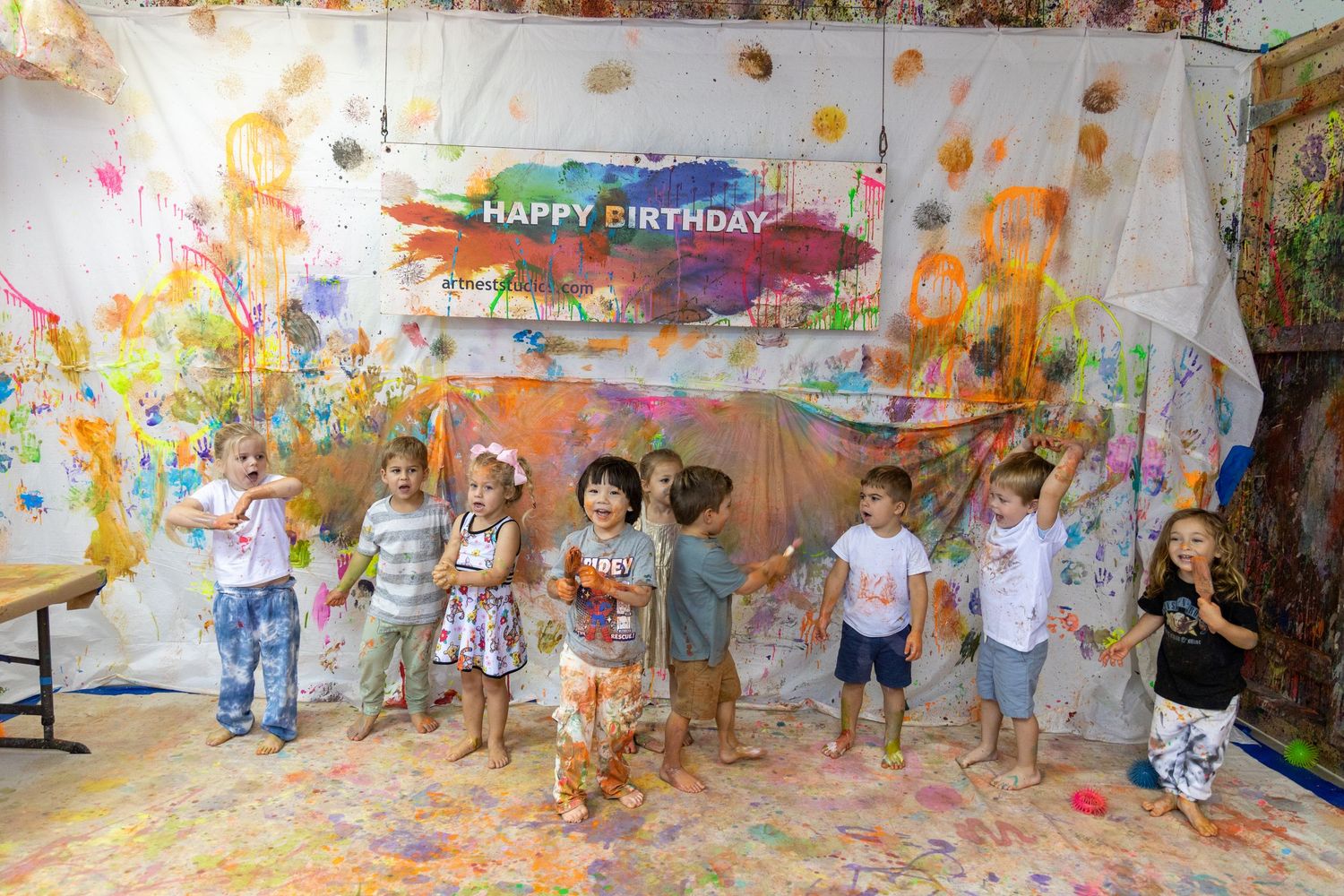 Children celebrating at a messy paint party with colorful splatters on walls and a Happy Birthday banner overhead.