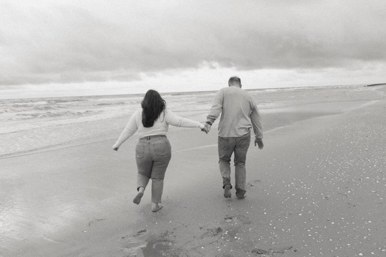 Black and white photo of couple running together along beach shoreline.