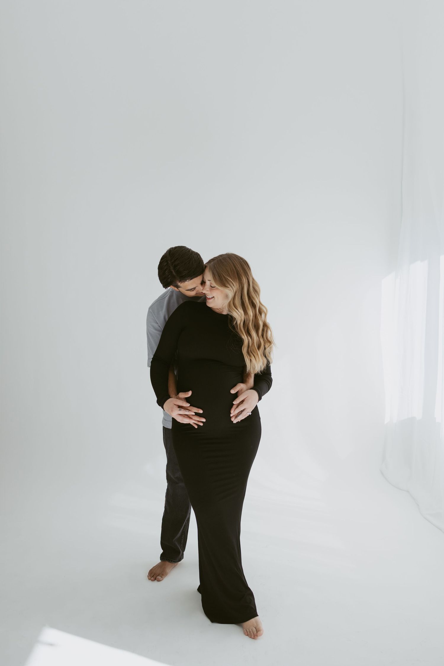 Intimate maternity portrait of couple embracing in black outfits against white background.