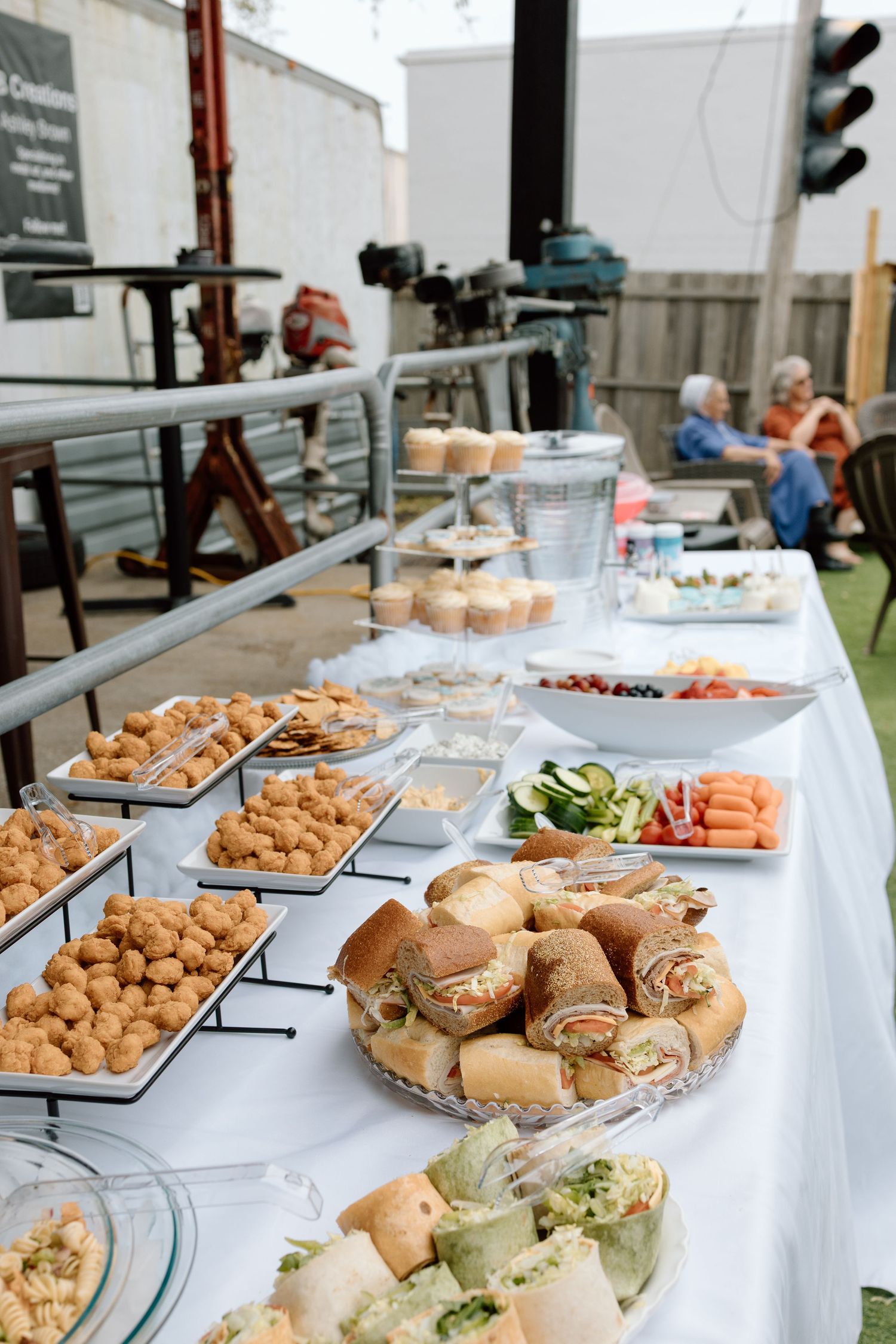 A catered buffet table with sandwiches, appetizers and various party foods spread on white linens at an outdoor event.