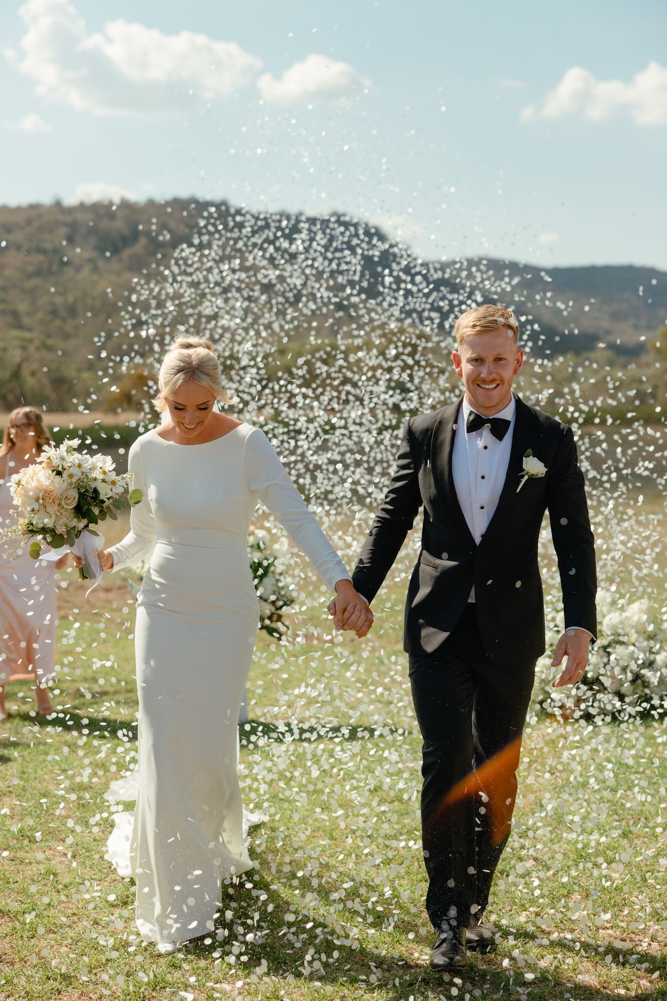 Newlyweds walking hand in hand through a shower of white confetti with mountains in the background.