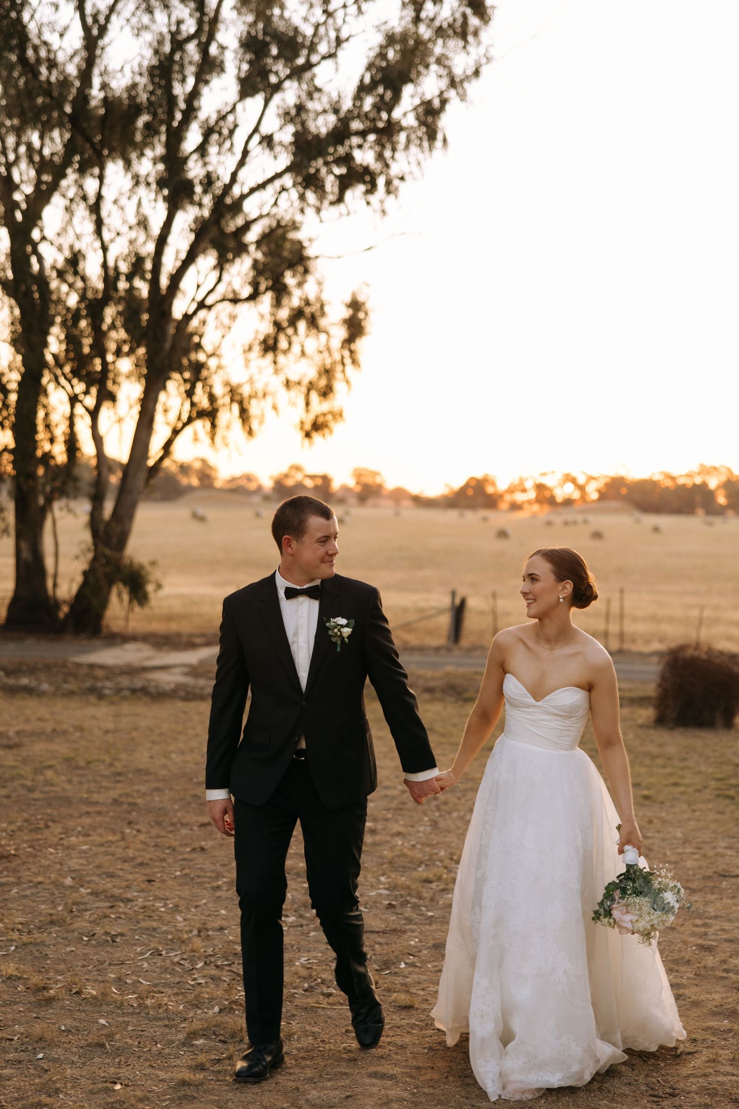 Couple walking together at sunset in formal wedding attire against rustic backdrop.