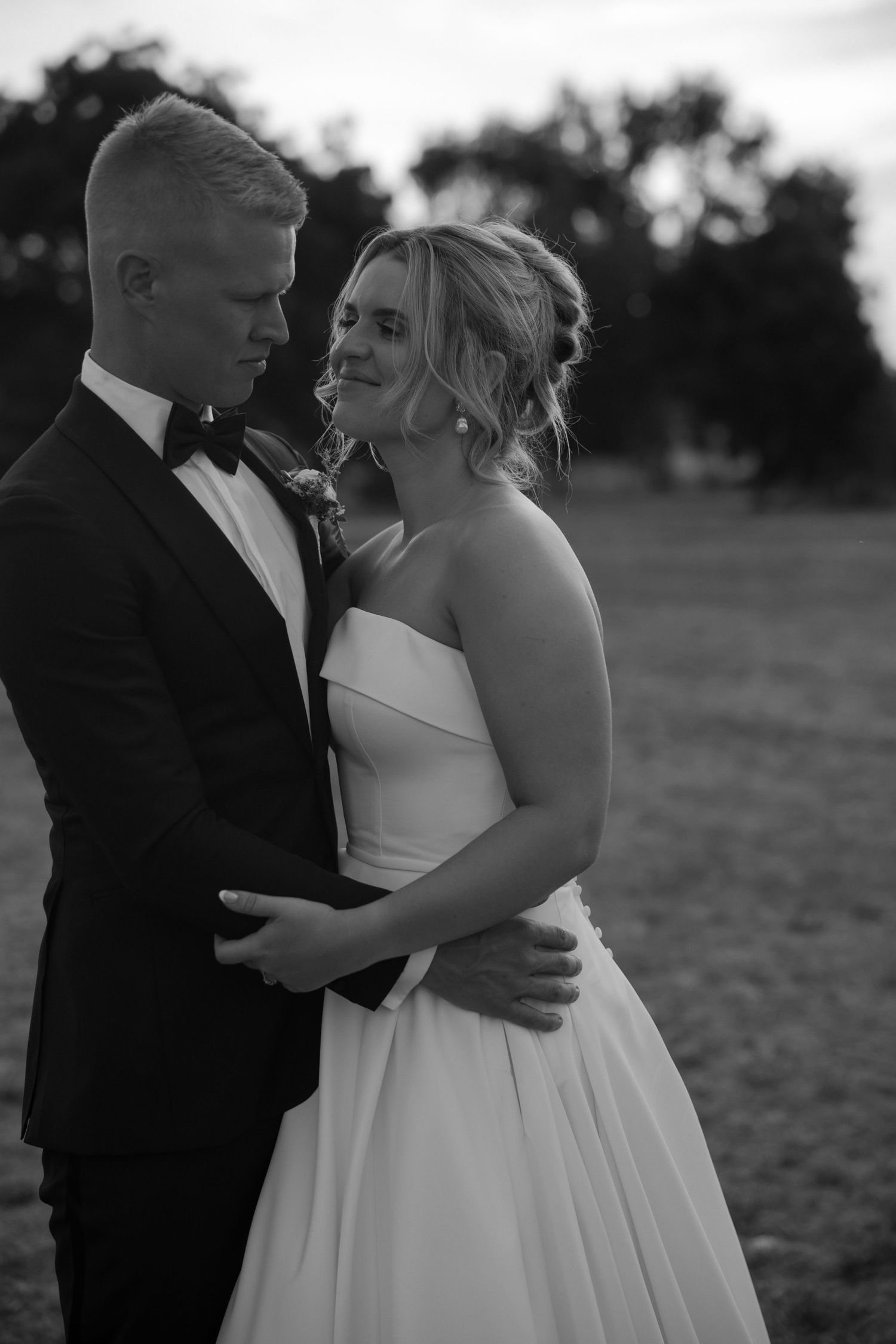 A romantic black and white wedding portrait of newlyweds embracing outdoors at dusk in formal attire.