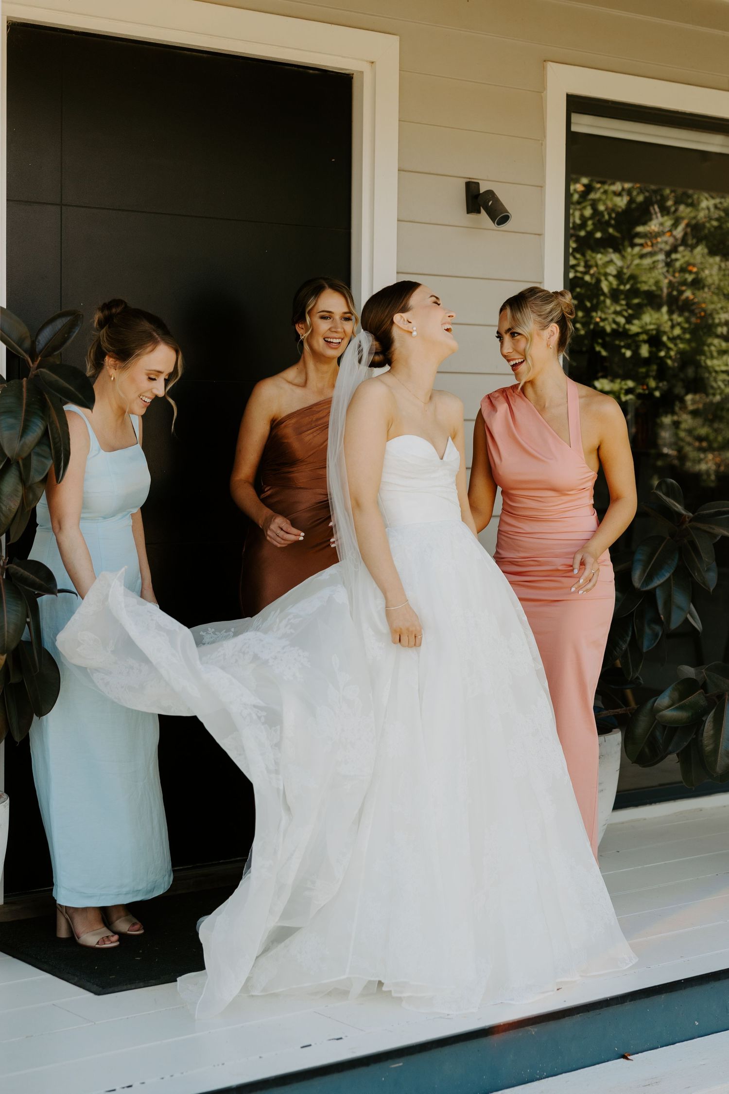 A group laughing together on a porch, wearing colorful bridesmaid dresses in blue, brown and pink around a white wedding gown.