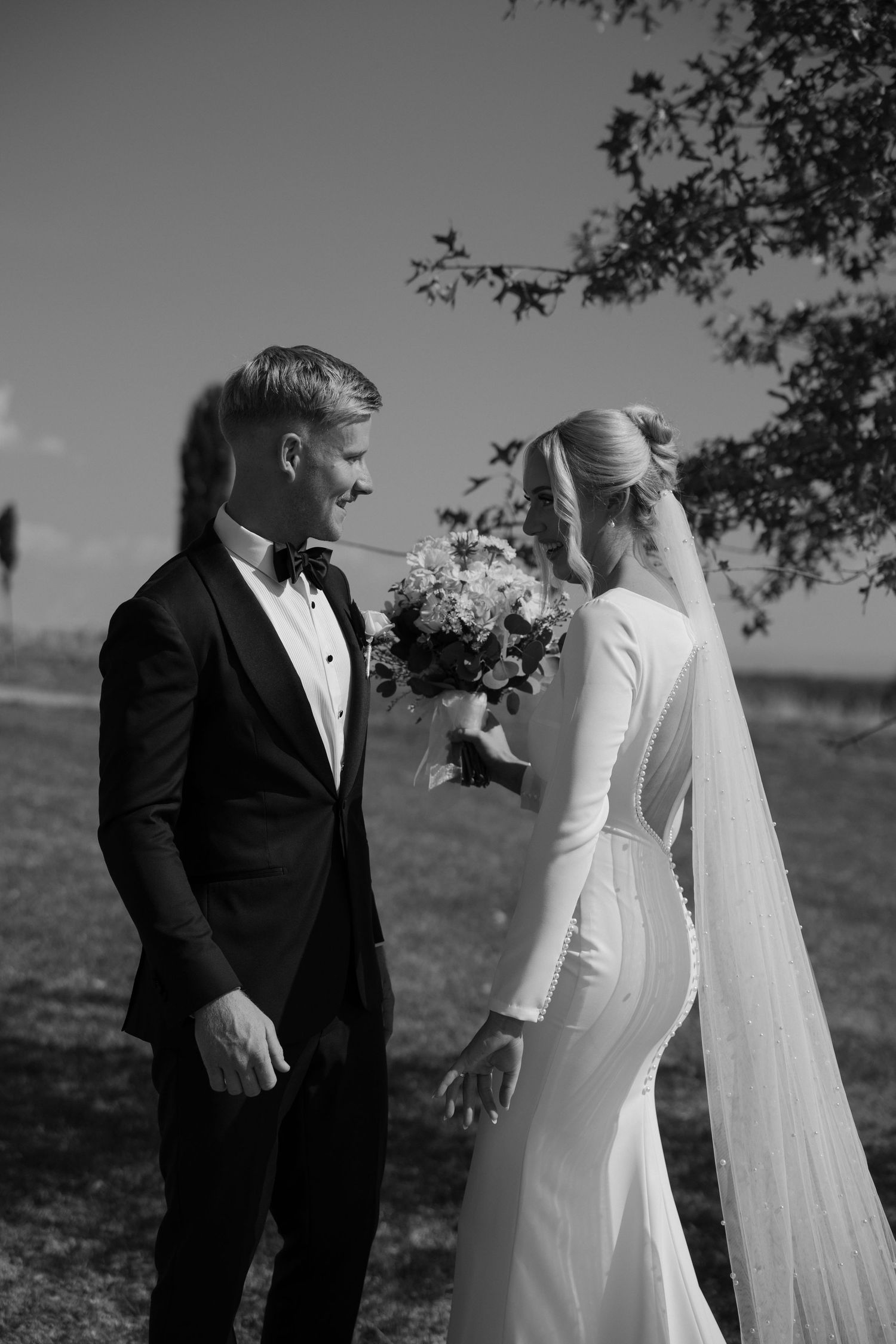 Black and white wedding photo capturing an elegant bride in a long-sleeved gown and veil with groom in tuxedo outdoors.