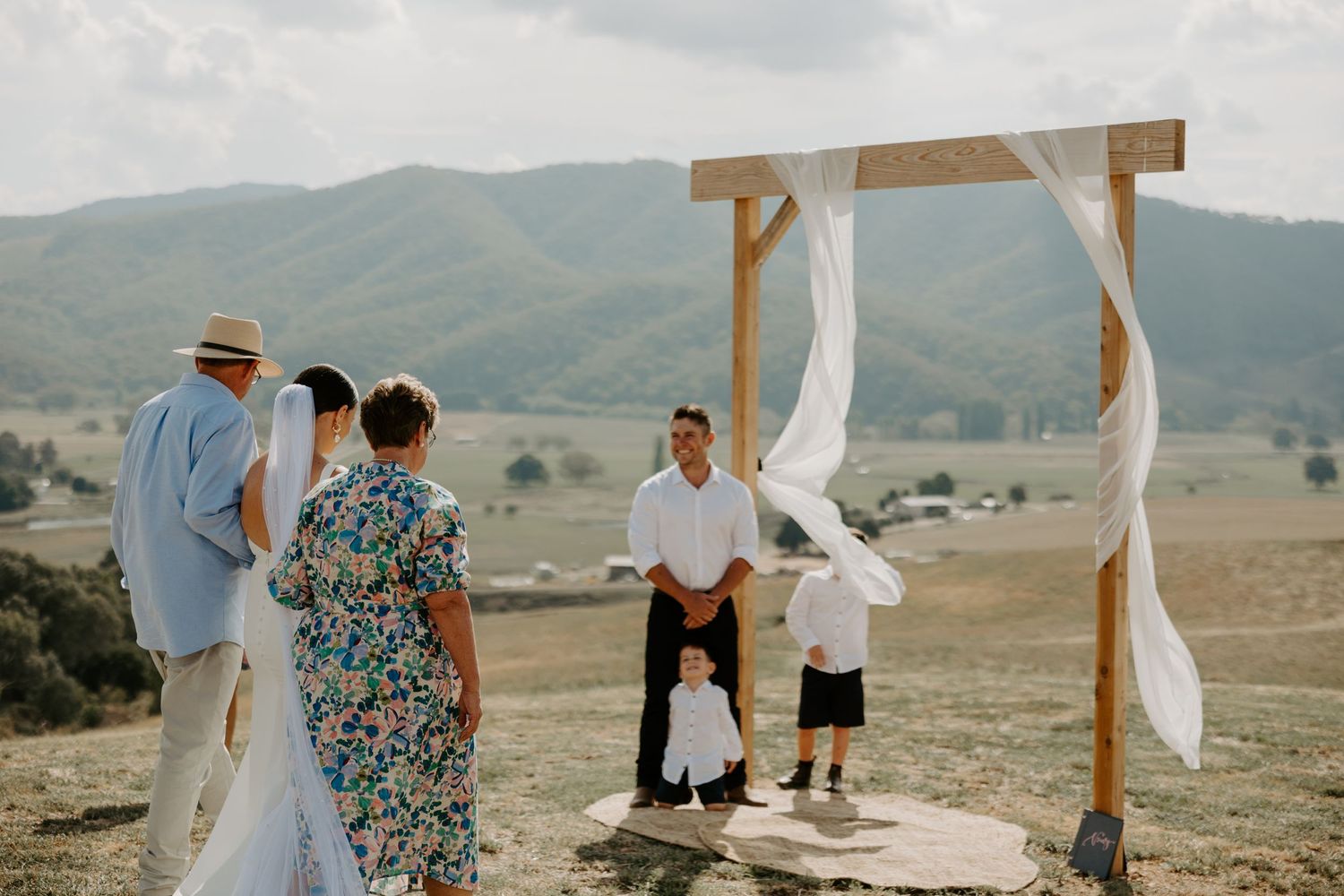 Wedding ceremony setup on hilltop with wooden arch and mountain views.