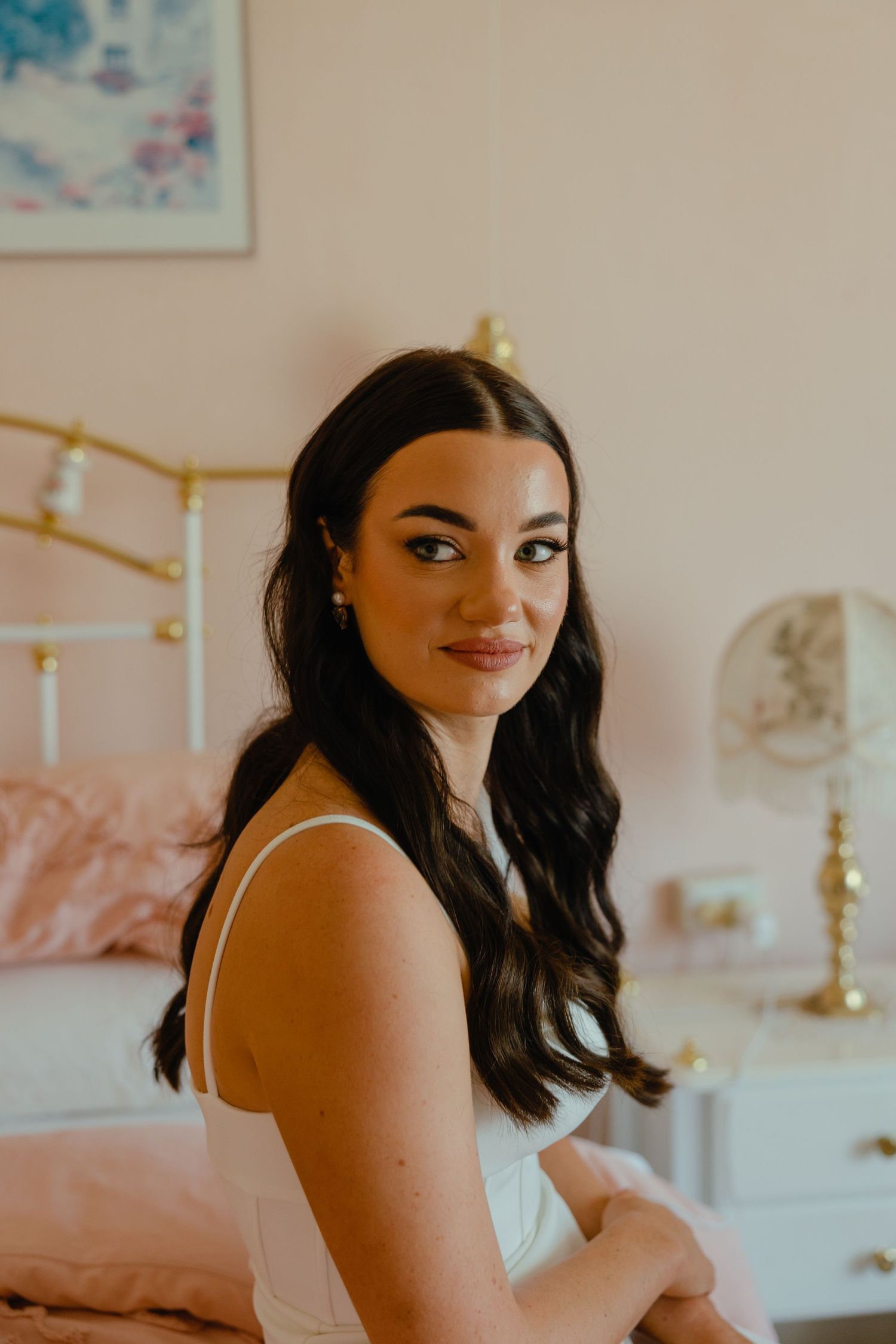 Bride in white dress sitting in bedroom with pink decor and vintage brass bed frame.