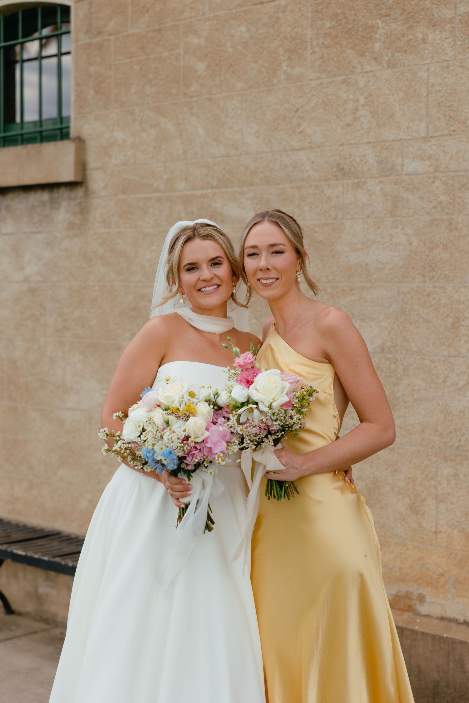Two wedding party members pose together, one in white bridal gown and one in yellow bridesmaid dress with floral bouquets.