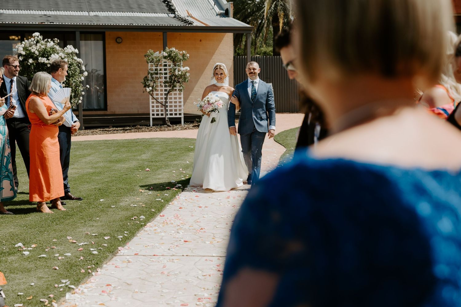 Outdoor garden wedding ceremony with floral arch decorations and guests gathered on a paved walkway.