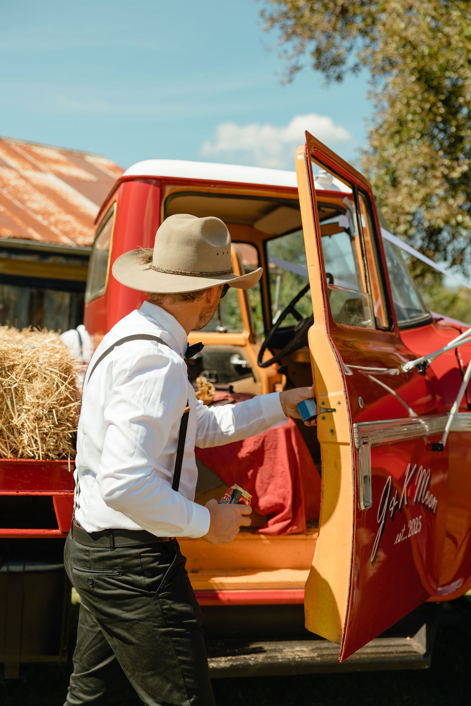 Person in cowboy hat and white shirt opening door of vintage red pickup truck with hay bales.