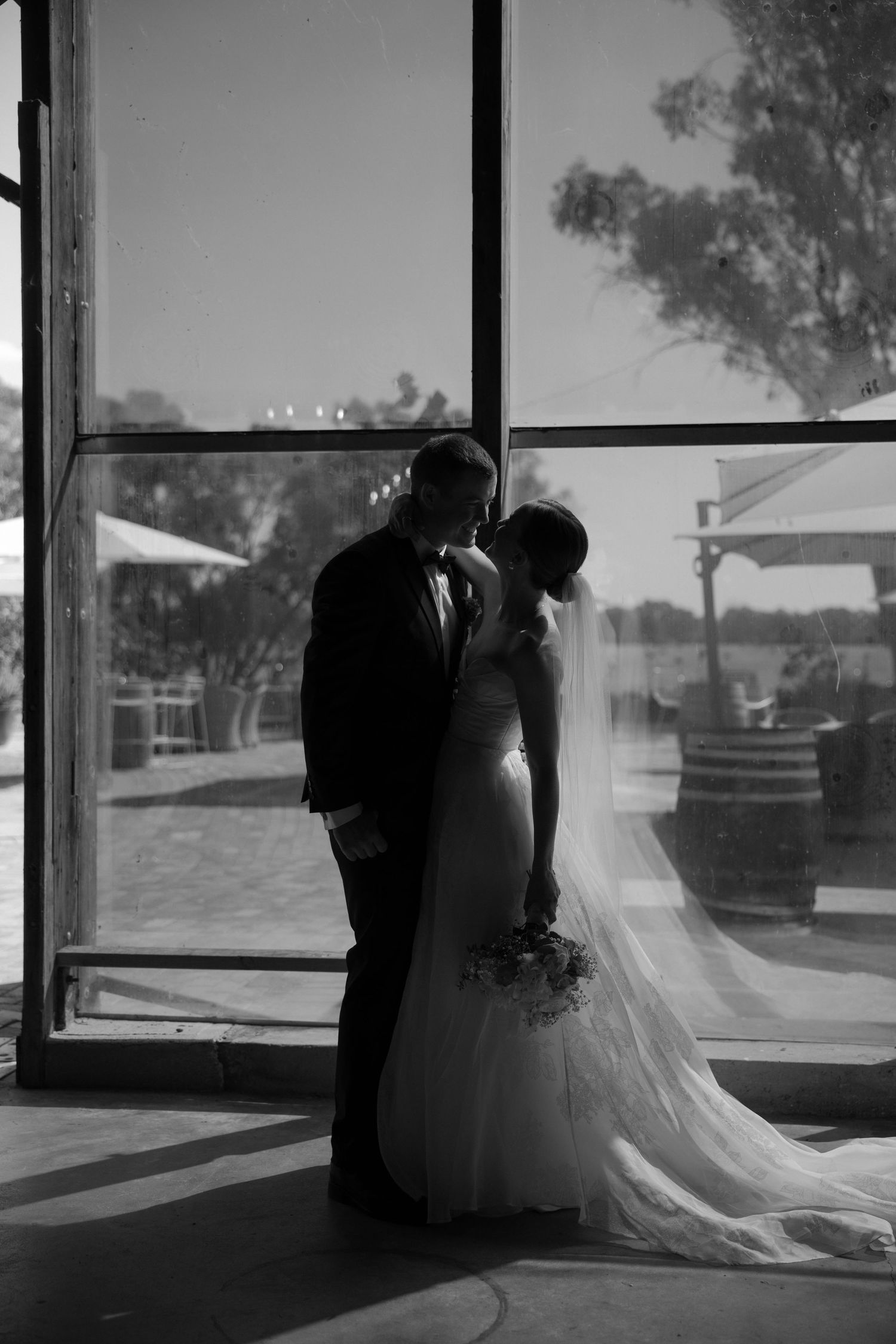 A silhouetted couple stands in front of a bright window, the bride in a flowing white gown and veil on their wedding day.