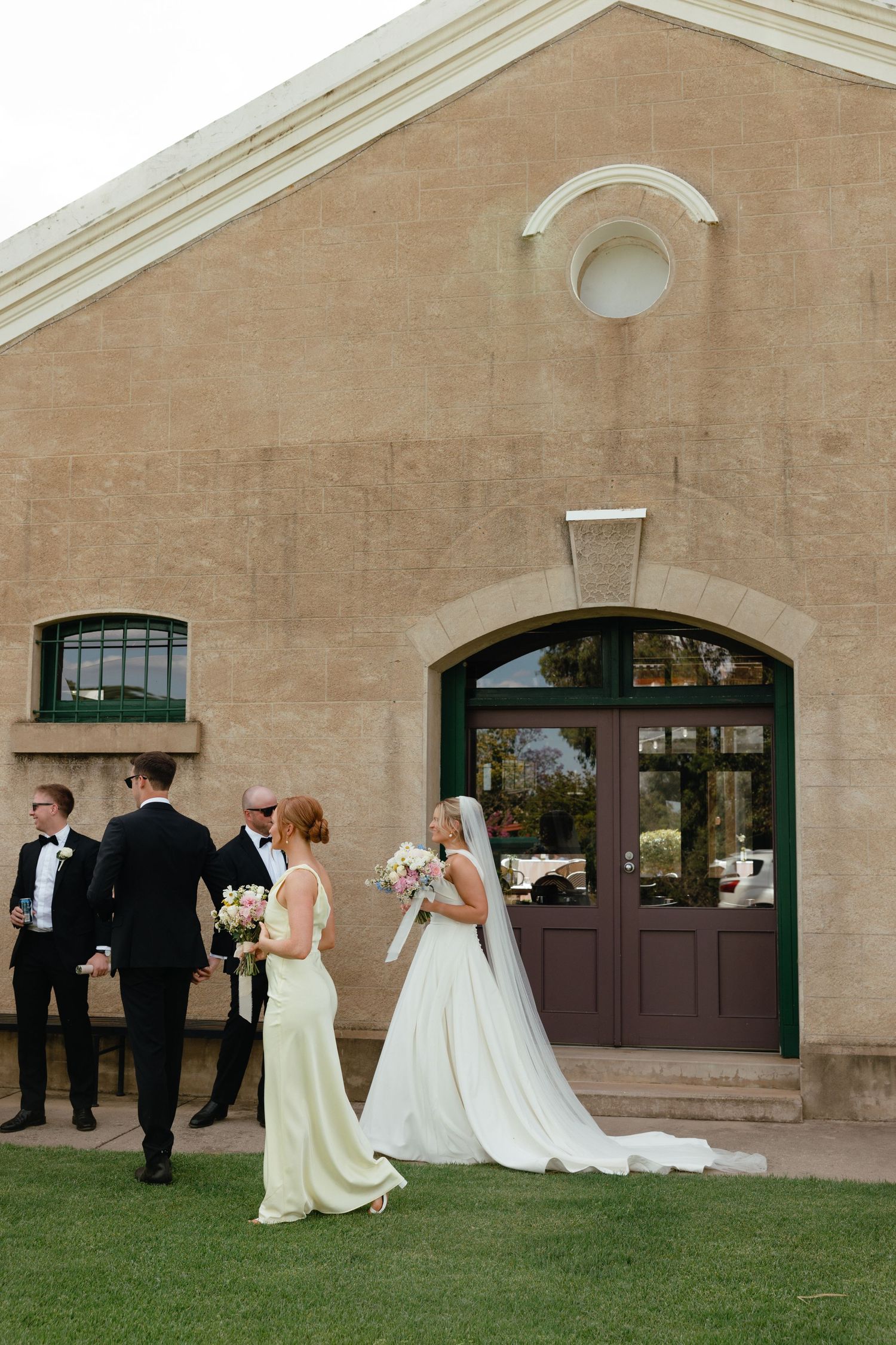 Wedding party gathered outside elegant stone building with arched doorway and manicured lawn.