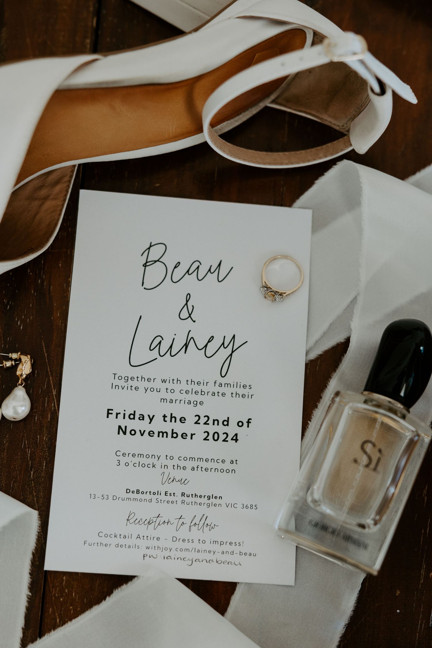 Wedding invitation with ring and perfume bottle on wooden surface displaying names Beau and Lainey for November 2024 date.