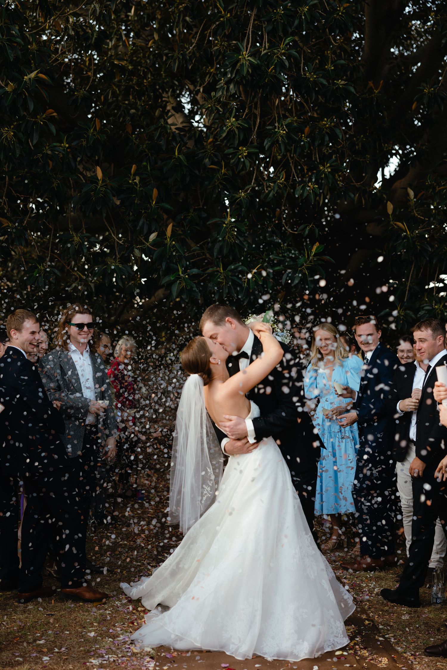 Newlyweds share a kiss under falling confetti surrounded by wedding guests.