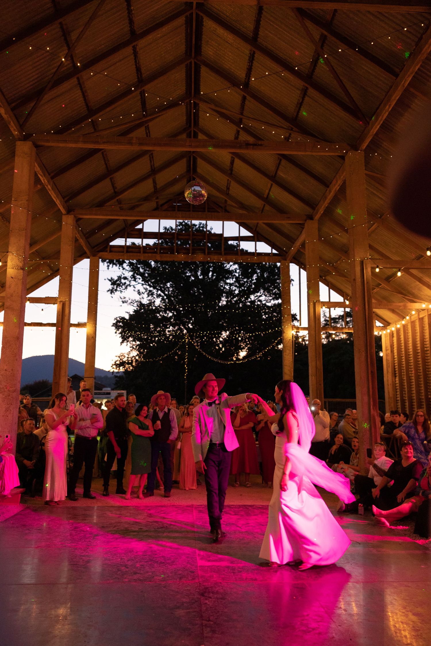 Wedding reception dancers glide across a wooden floor under a rustic barn roof illuminated by pink lighting and string lights.