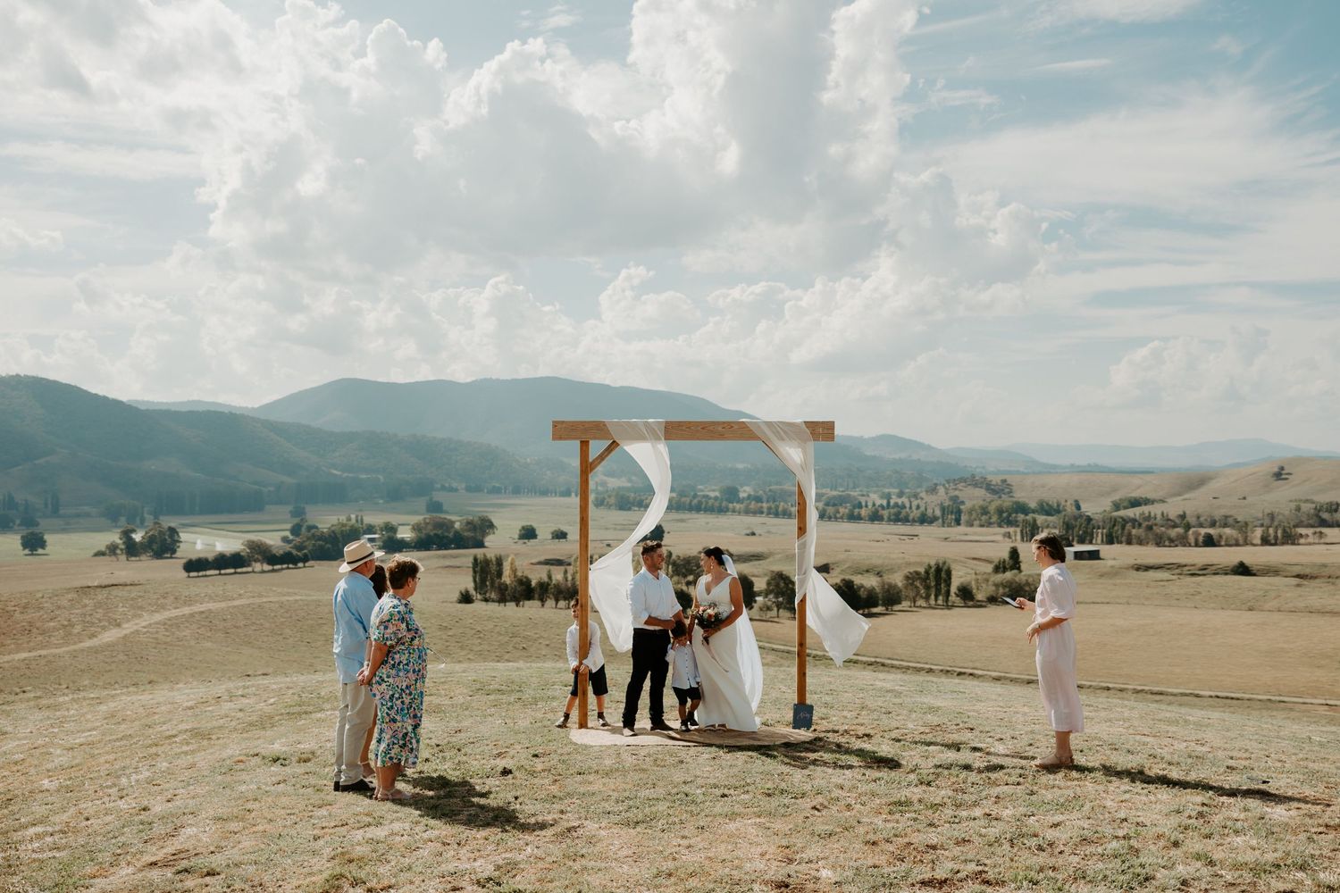 Outdoor wedding ceremony under wooden arch with white fabric and mountain landscape backdrop.