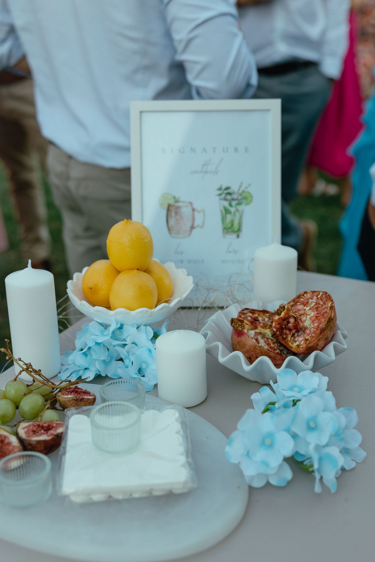 Table decorated with fresh citrus fruits, blue hydrangeas, white candles, and a framed menu sign for an elegant event setting.