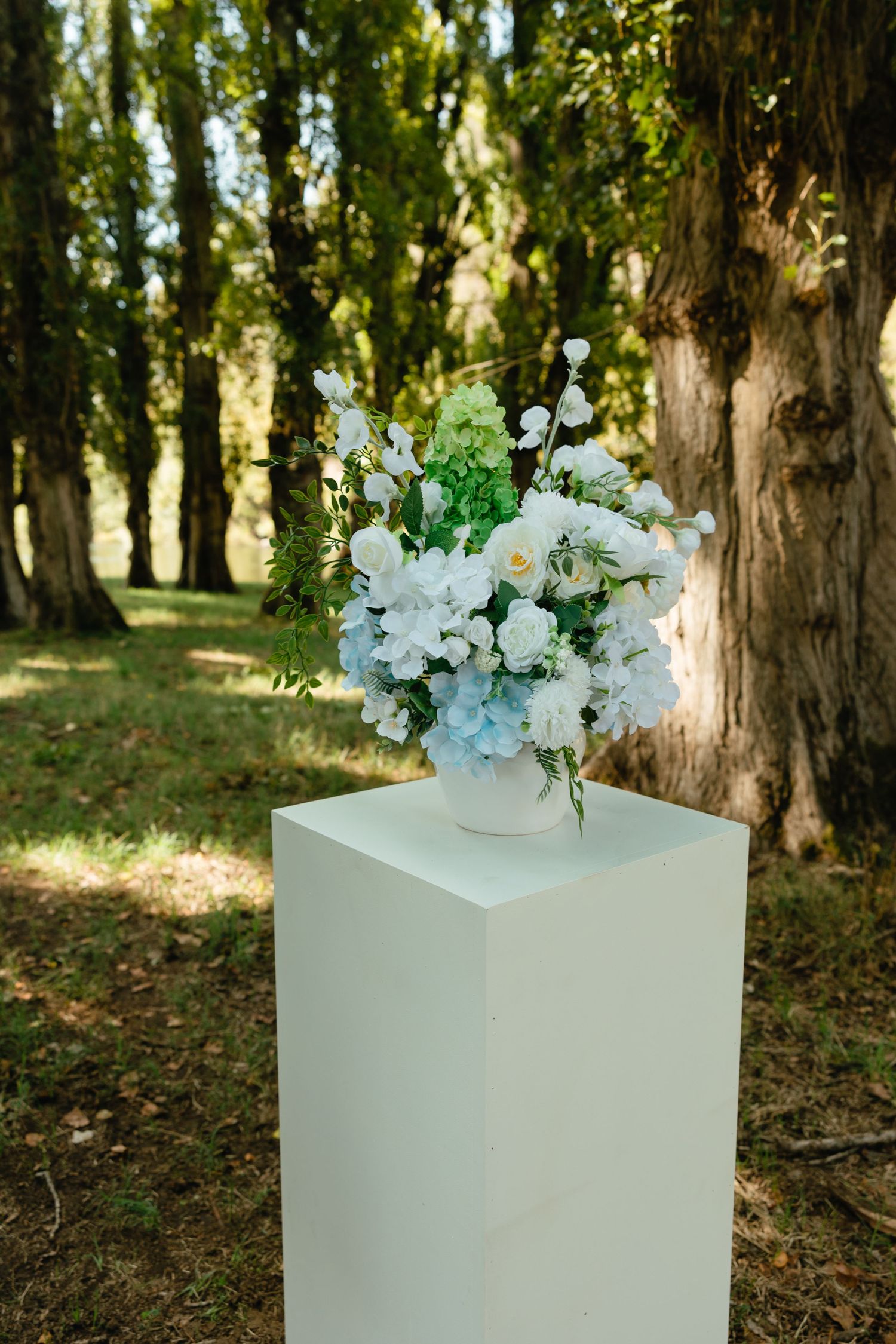 Elegant white floral arrangement in a vase displayed on a tall white pedestal column outdoors among trees in natural light.